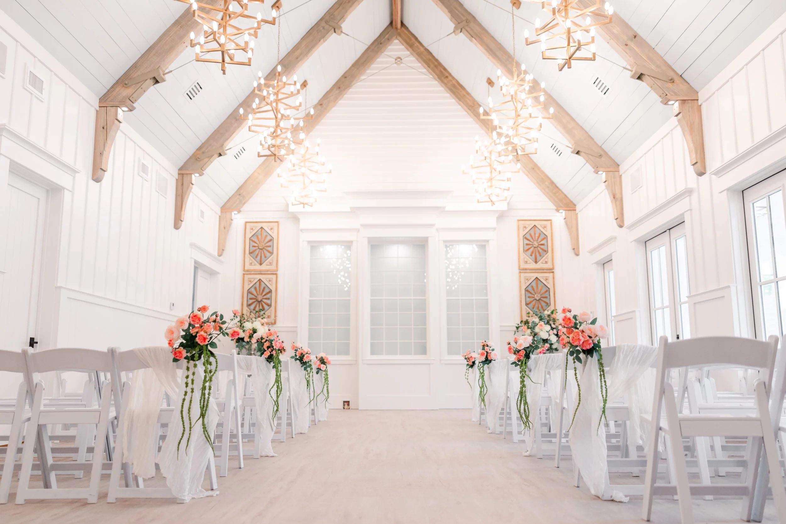 Elegant wedding venue with white chairs, floral decorations, and modern chandeliers.