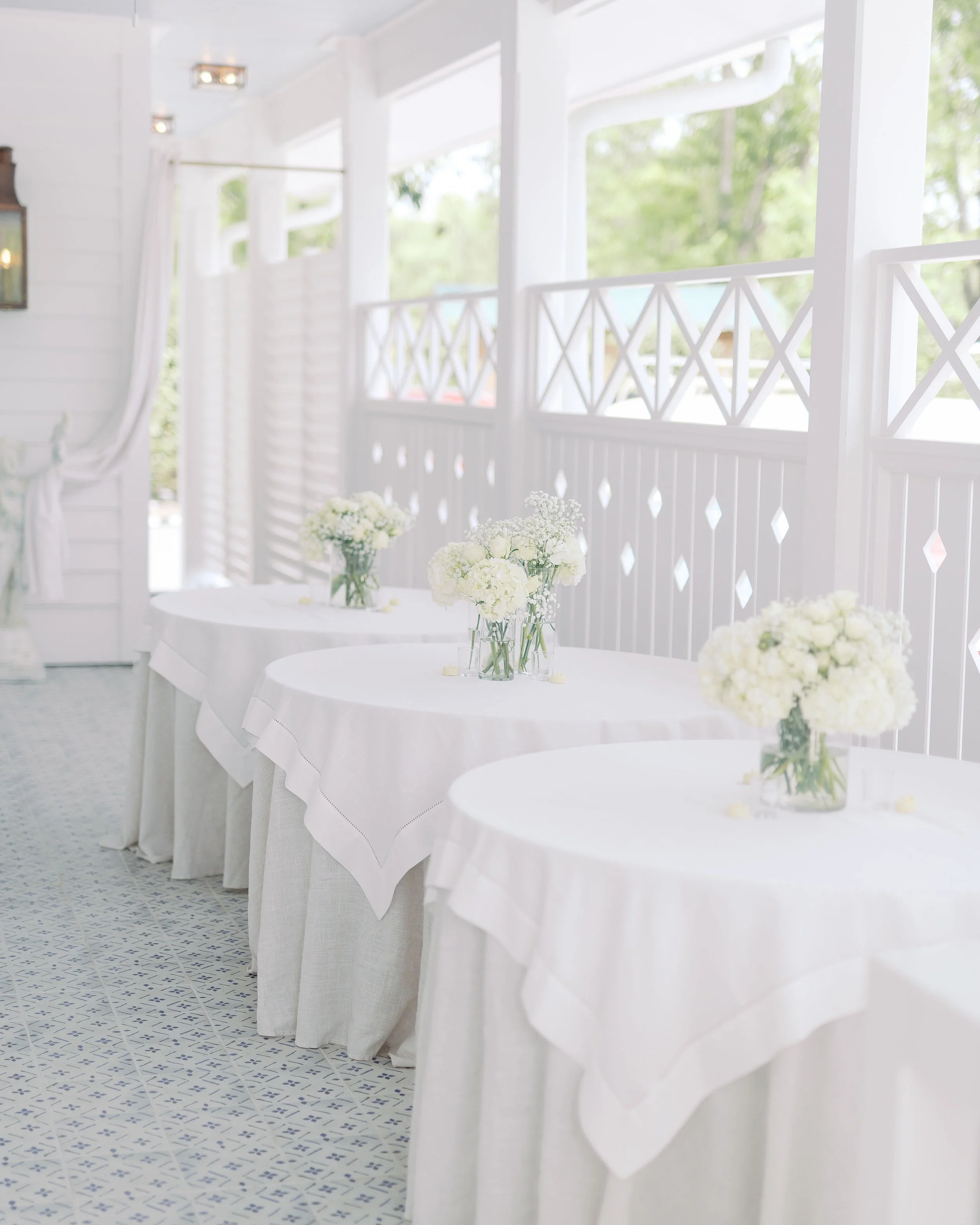 Three round tables with white tablecloths and vases of white flowers on a sunny porch with white railings and windows.