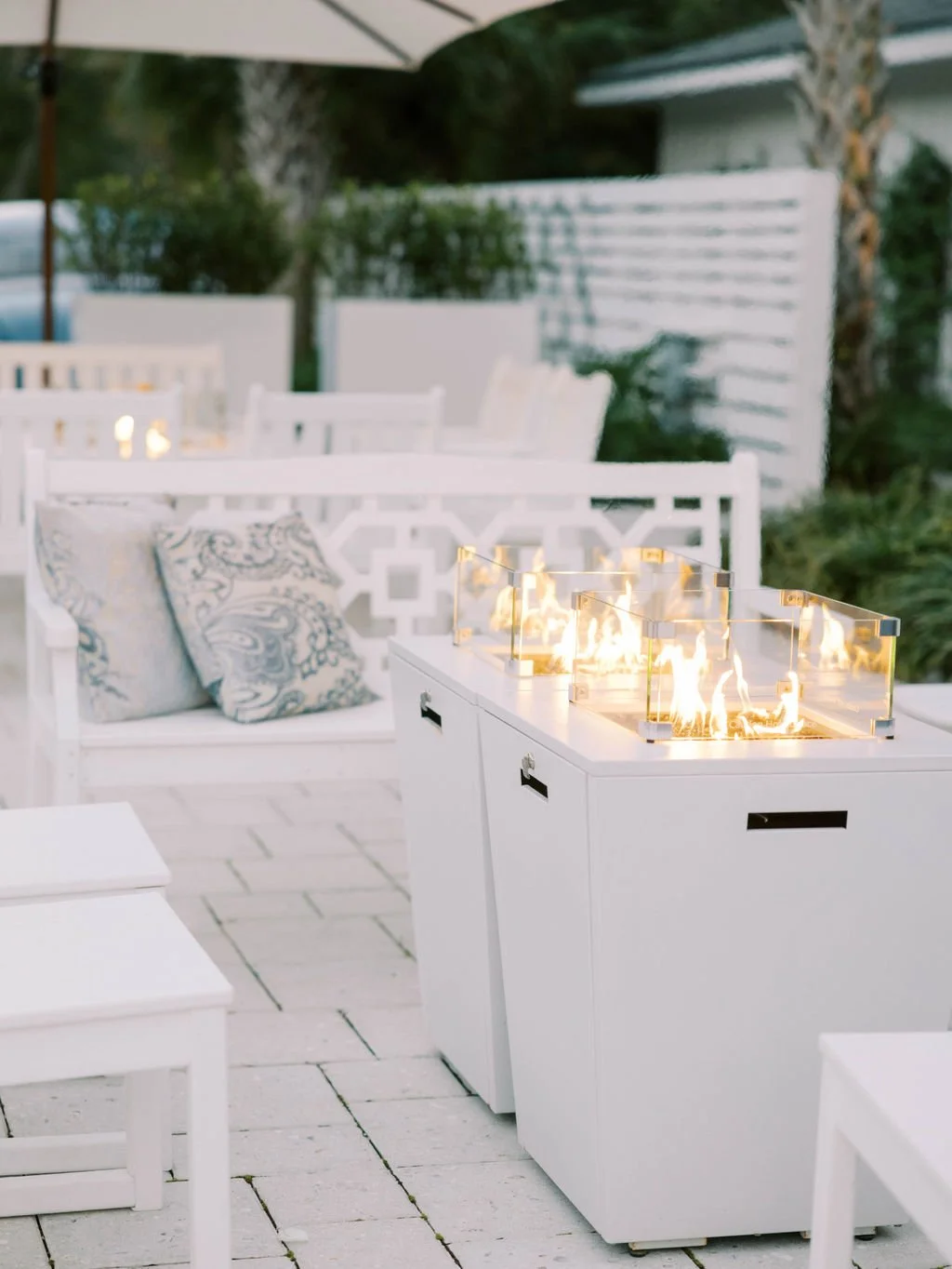 Outdoor patio with white furniture, decorative pillows, and a modern rectangular fire pit generating flames, surrounded by a brick floor and a white privacy fence in the background.