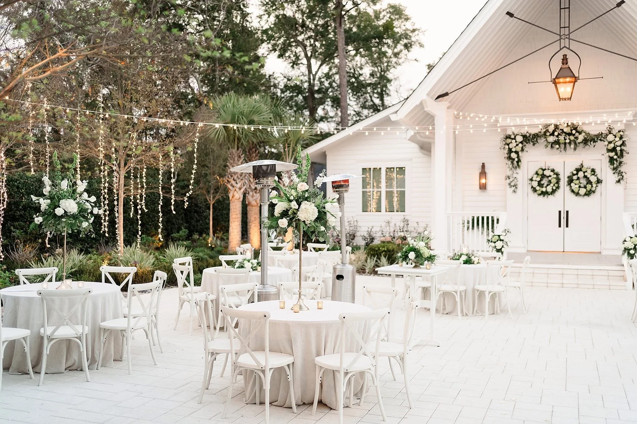 Outdoor wedding venue setup with white tables and chairs, floral centerpieces, string lights, and a decorated white building in the background.