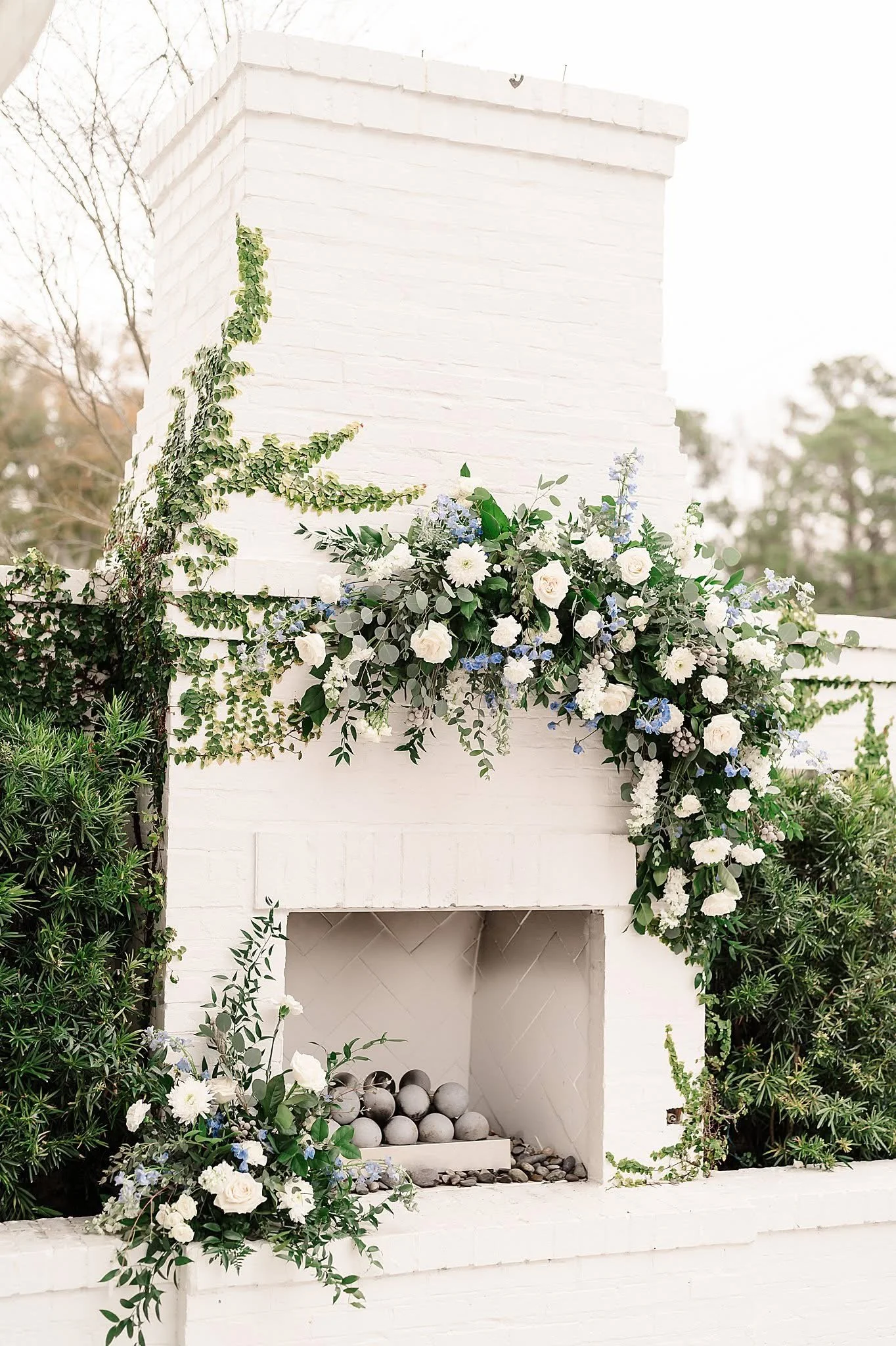 White brick outdoor fireplace adorned with white and blue floral arrangements and greenery.