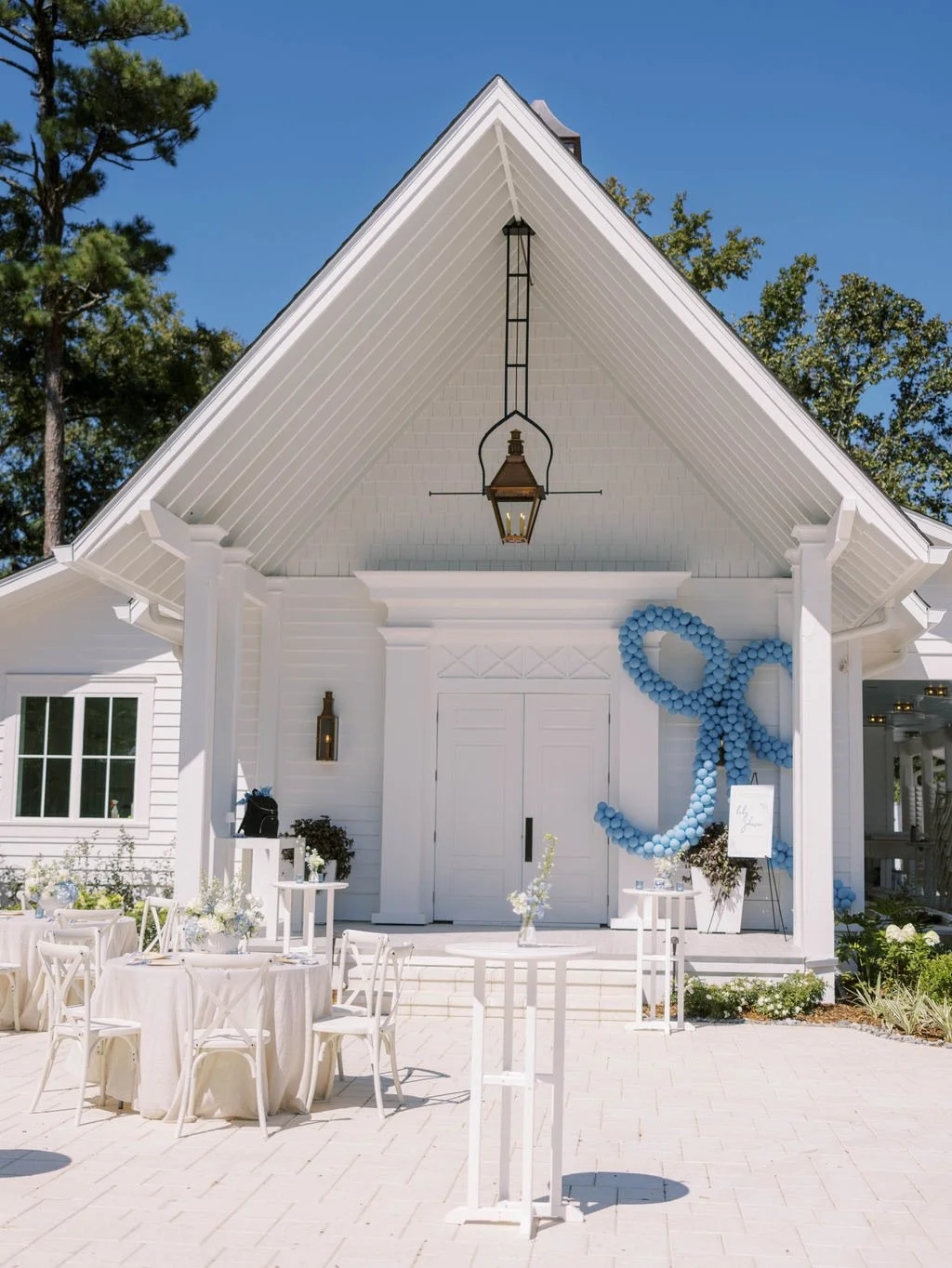 White building entrance with triangular roof and lantern. Blue balloon decoration shaped like a ribbon adorns the entrance. Tables with white tablecloths and chairs set up on patio.