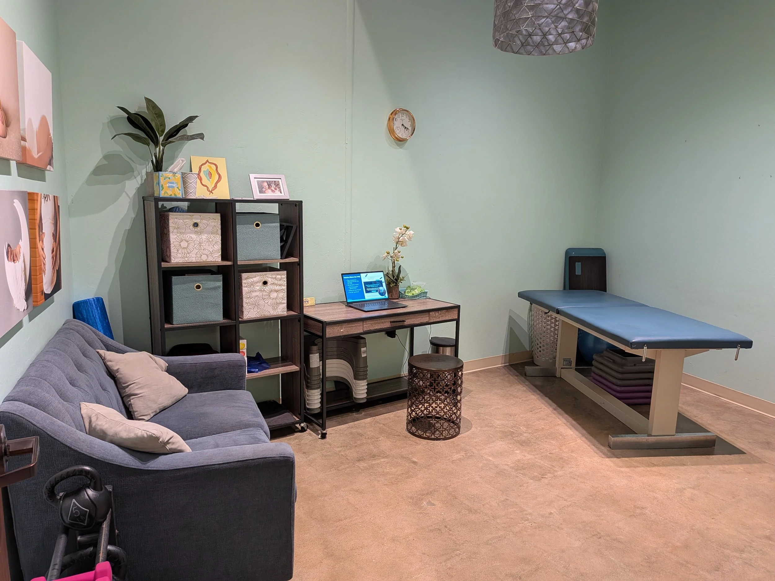 A therapy or examination room with a grey sofa with pillows, a black shelf filled with storage bins, a small table with a laptop and a flowered plant, a massage or examination table, and a light green wall with pictures and a clock.