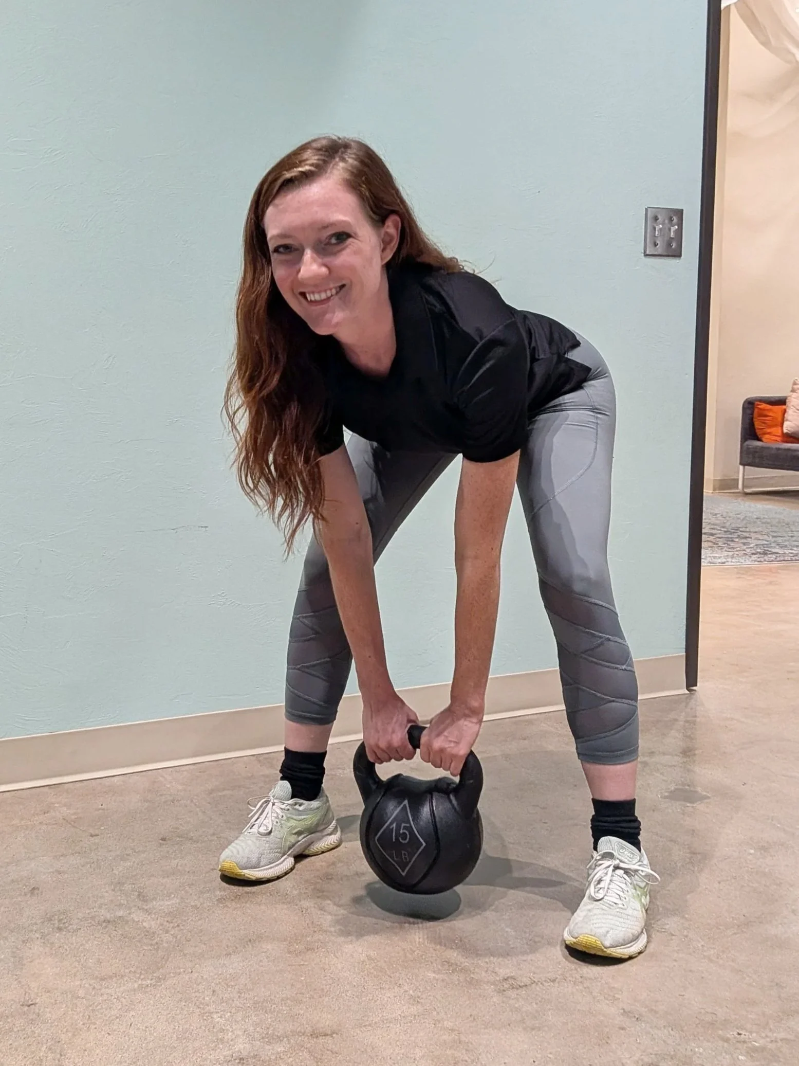 Woman with long red hair in athletic wear lifting a 15-pound kettlebell in an indoor gym or fitness area.
