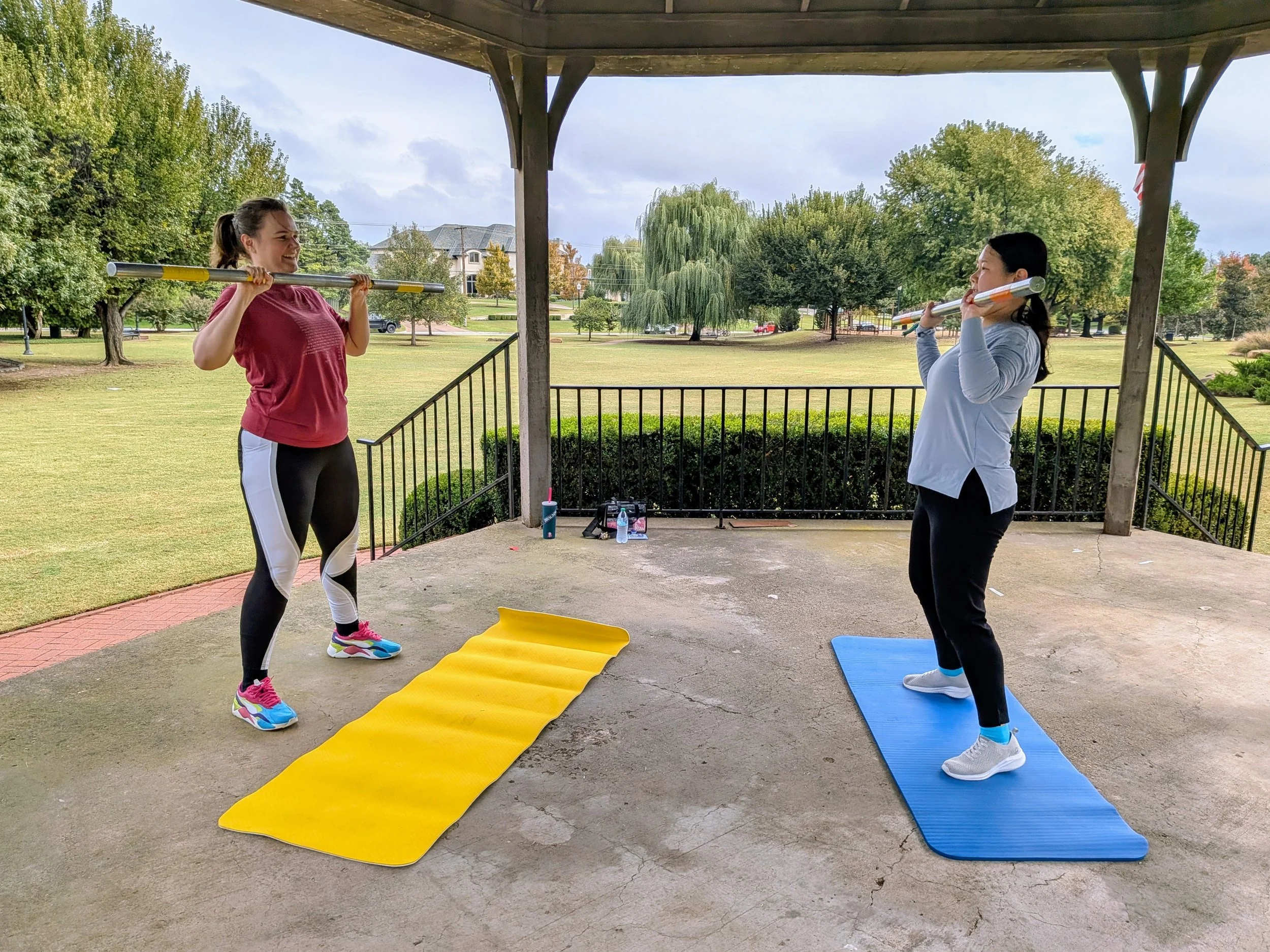 A photo of two people working out at a public park outside. They are holding squat bars and are standing in front of exercise yoga mats.