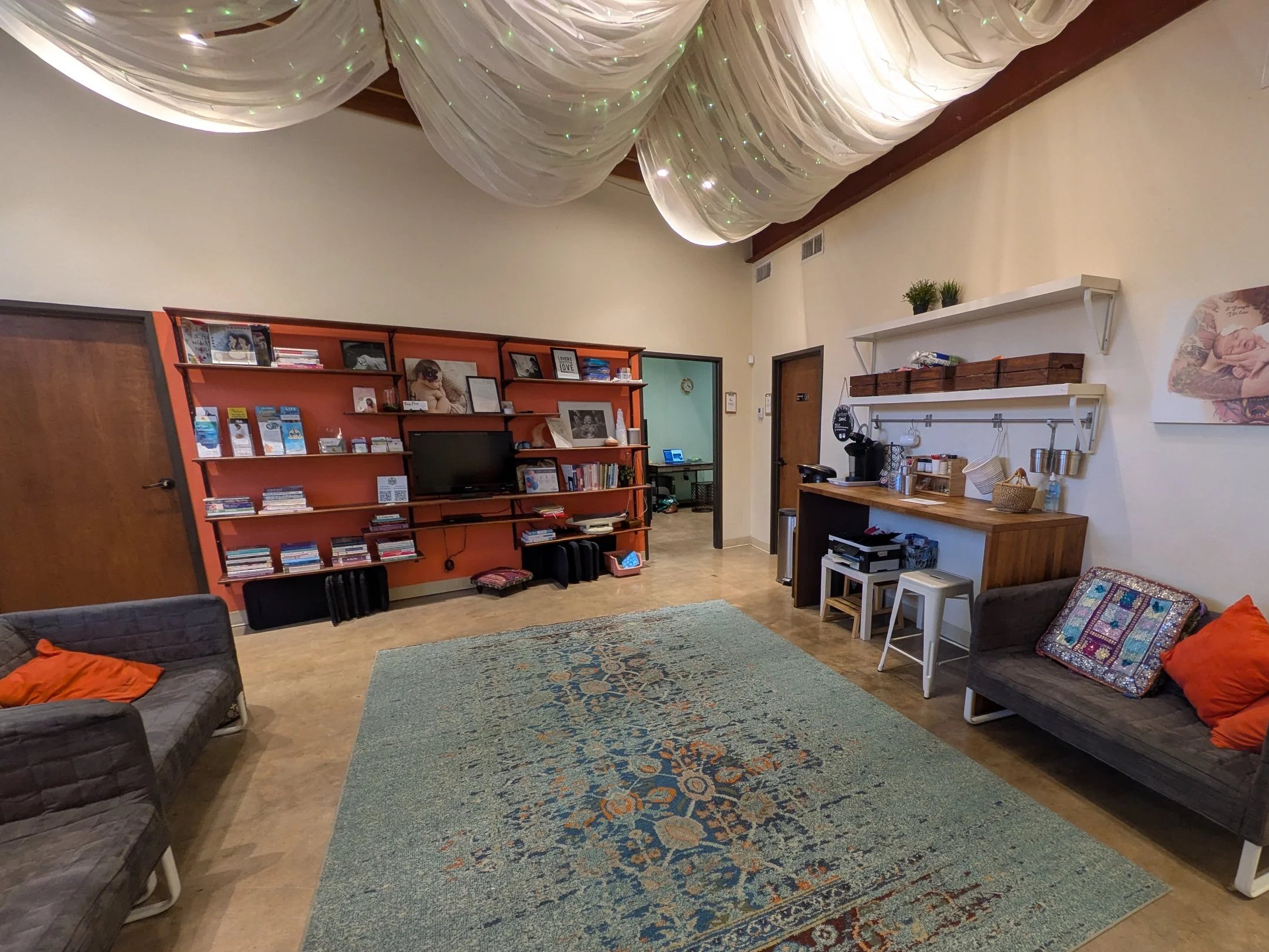 A cozy living room with a large vintage rug, two gray couches with orange pillows, a wooden bar counter, shelves with decorations, and a colorful draped ceiling with lights.