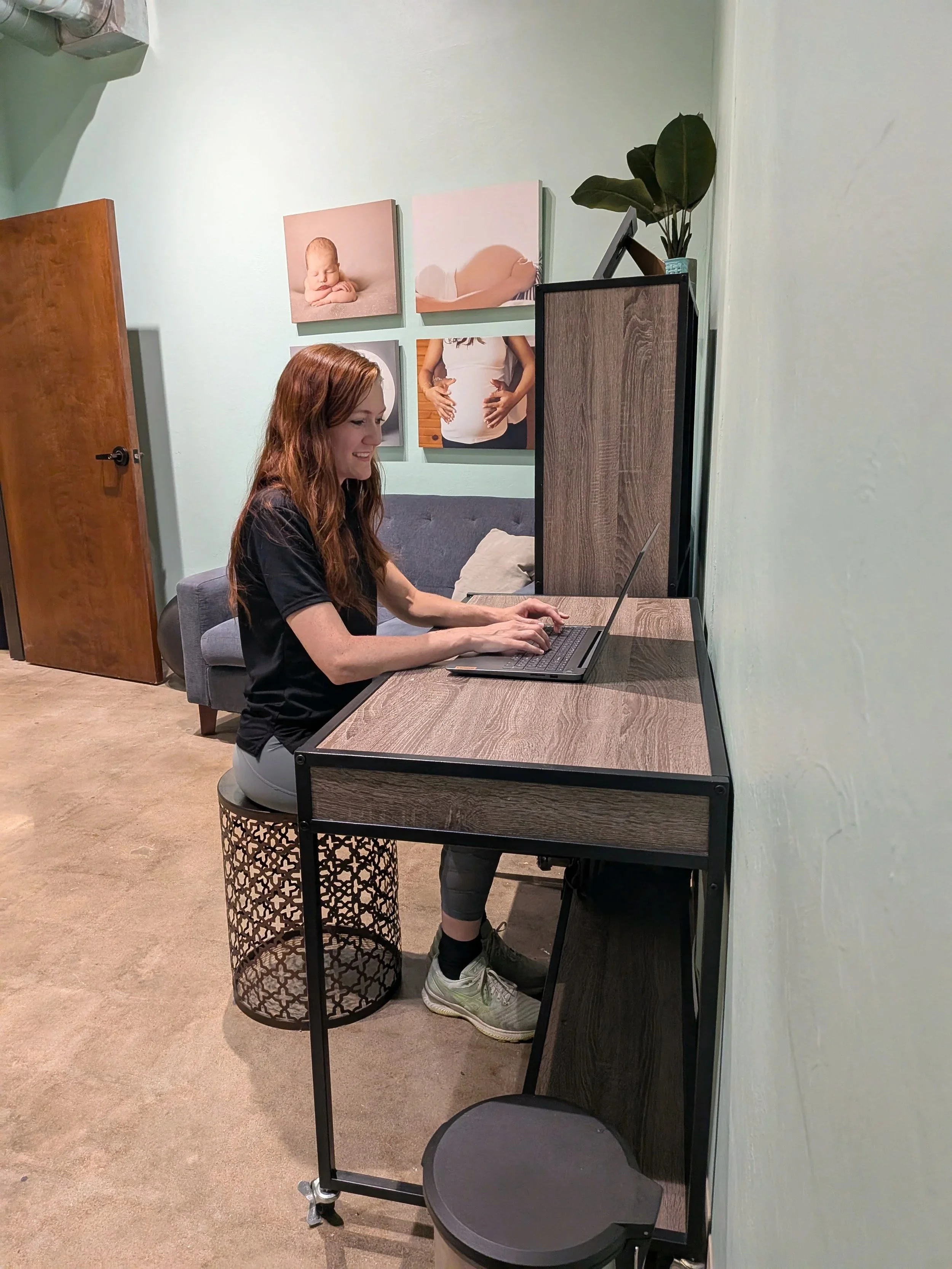 A woman with red hair sitting and smiling at a computer screen. There are pictures on the wall behind her related to pregnancy.