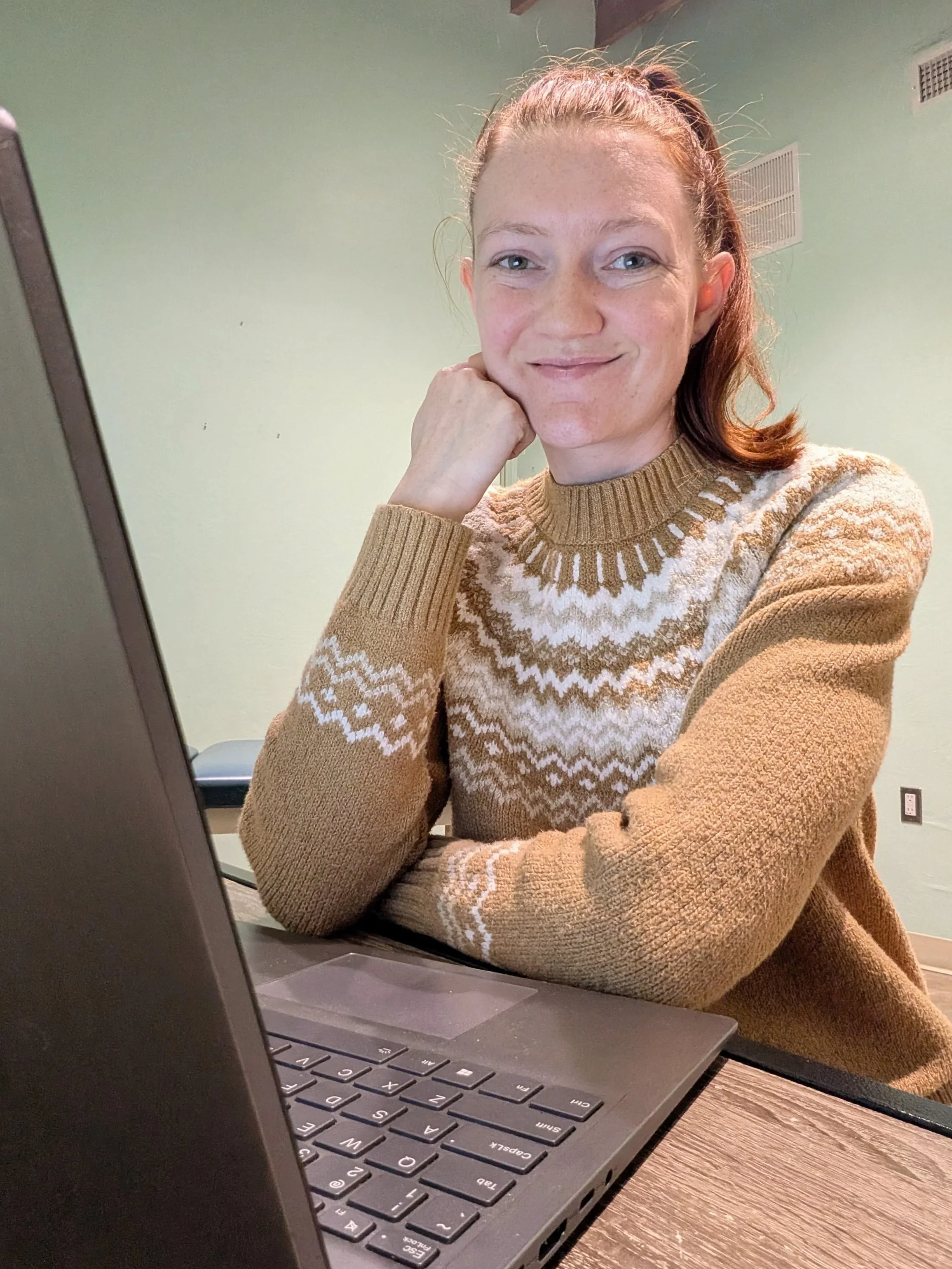 A woman with red hair pulled back, wearing a tan and white patterned sweater, sitting at a table with a laptop, resting her chin on her hand, smiling at the camera.