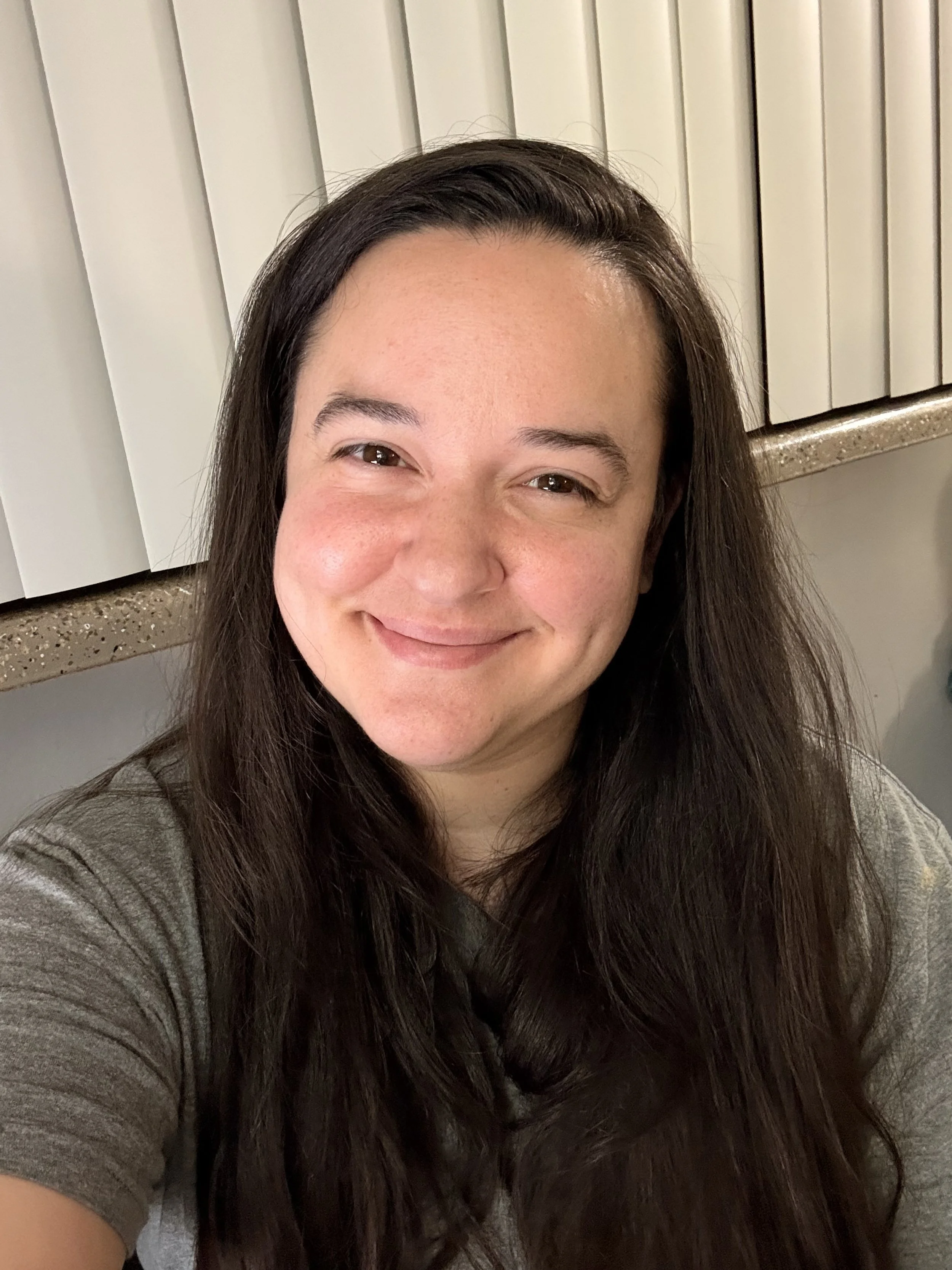 A woman with long dark hair smiling at the camera in front of vertical blinds.