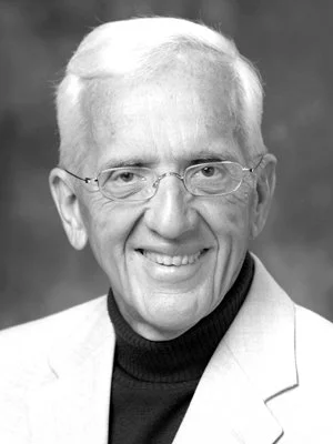 Black and white portrait of an elderly man smiling, wearing glasses, a dark turtleneck, and a light-colored blazer.