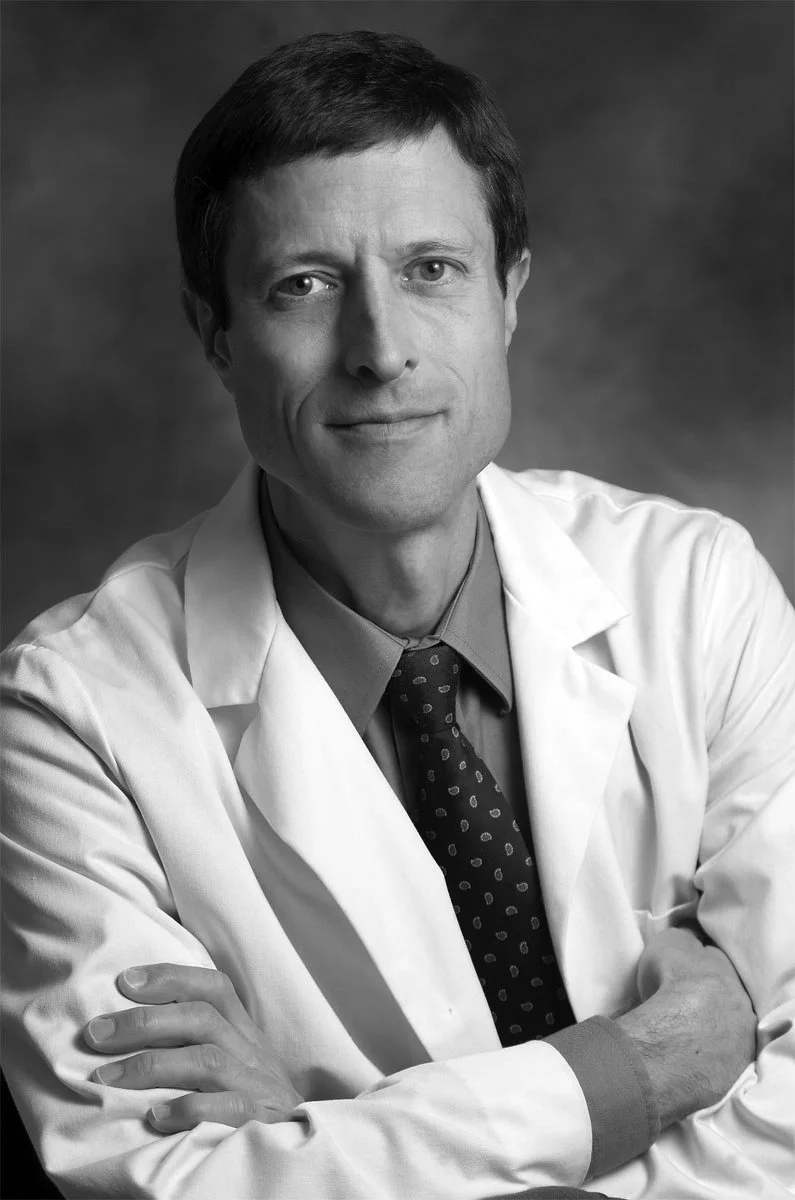 Black and white photo of a middle-aged man wearing a white lab coat, shirt, and tie, with arms crossed, looking confidently at the camera.