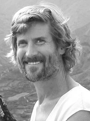 Black and white photo of a smiling man with wavy hair and a beard, outdoors with a mountainous background.