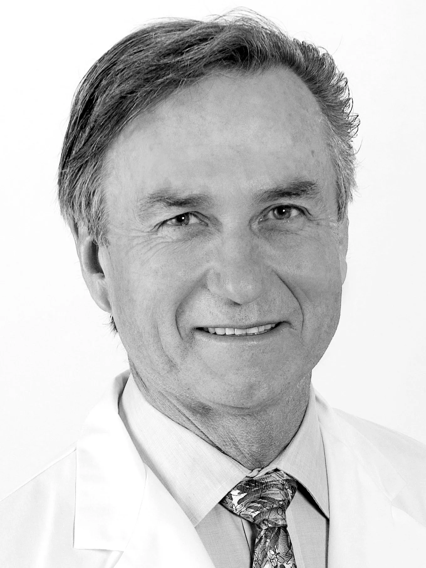 Black and white portrait of a middle-aged man with light hair, wearing a white shirt and patterned tie, smiling slightly.