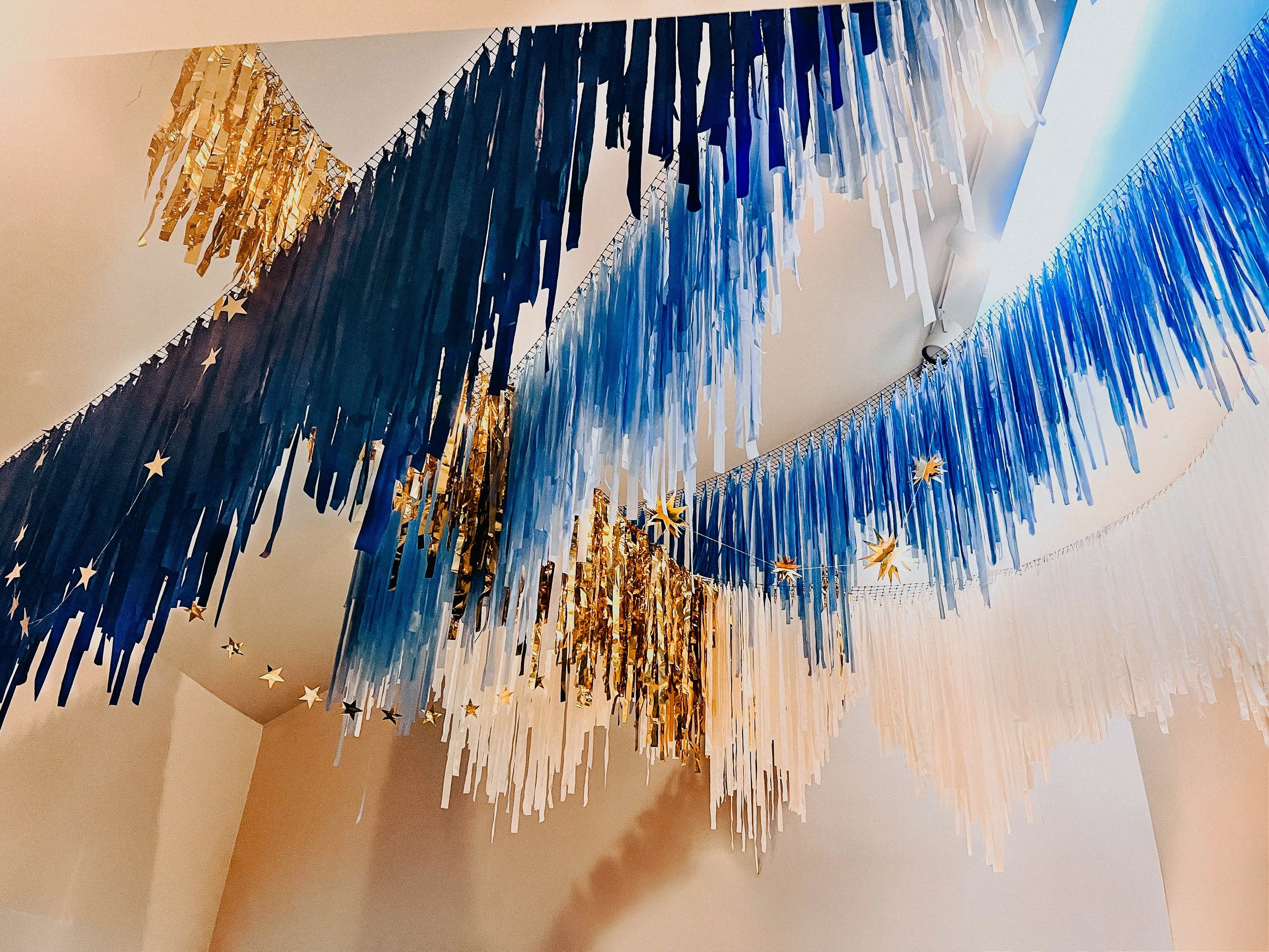 Colorful hanging decorations in blues, gold, and white, with star shapes, strung across a ceiling. Aerial fringe installation.