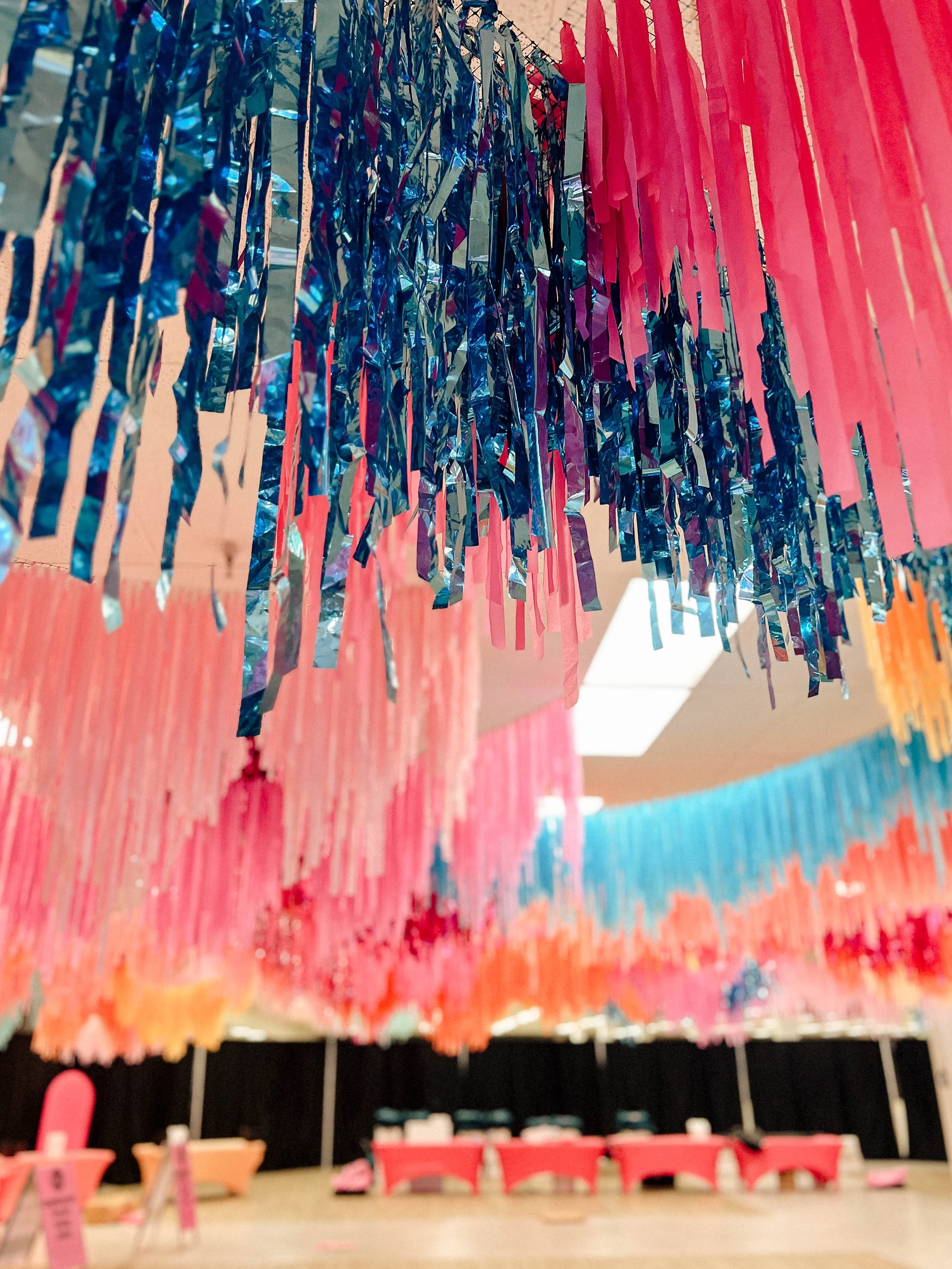 Decorative pink and blue aerial fringe instalation in an indoor setting, with a table featuring small pink and red items and chairs in the background.