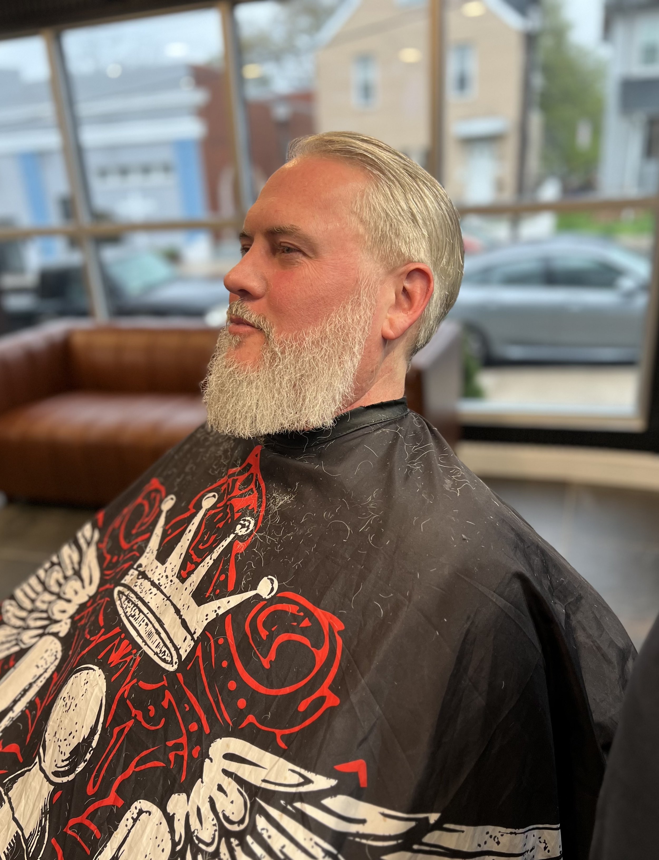 Man with a gray beard and slicked-back gray hair getting a haircut in a salon, wearing a black cape with red and white graphic design.