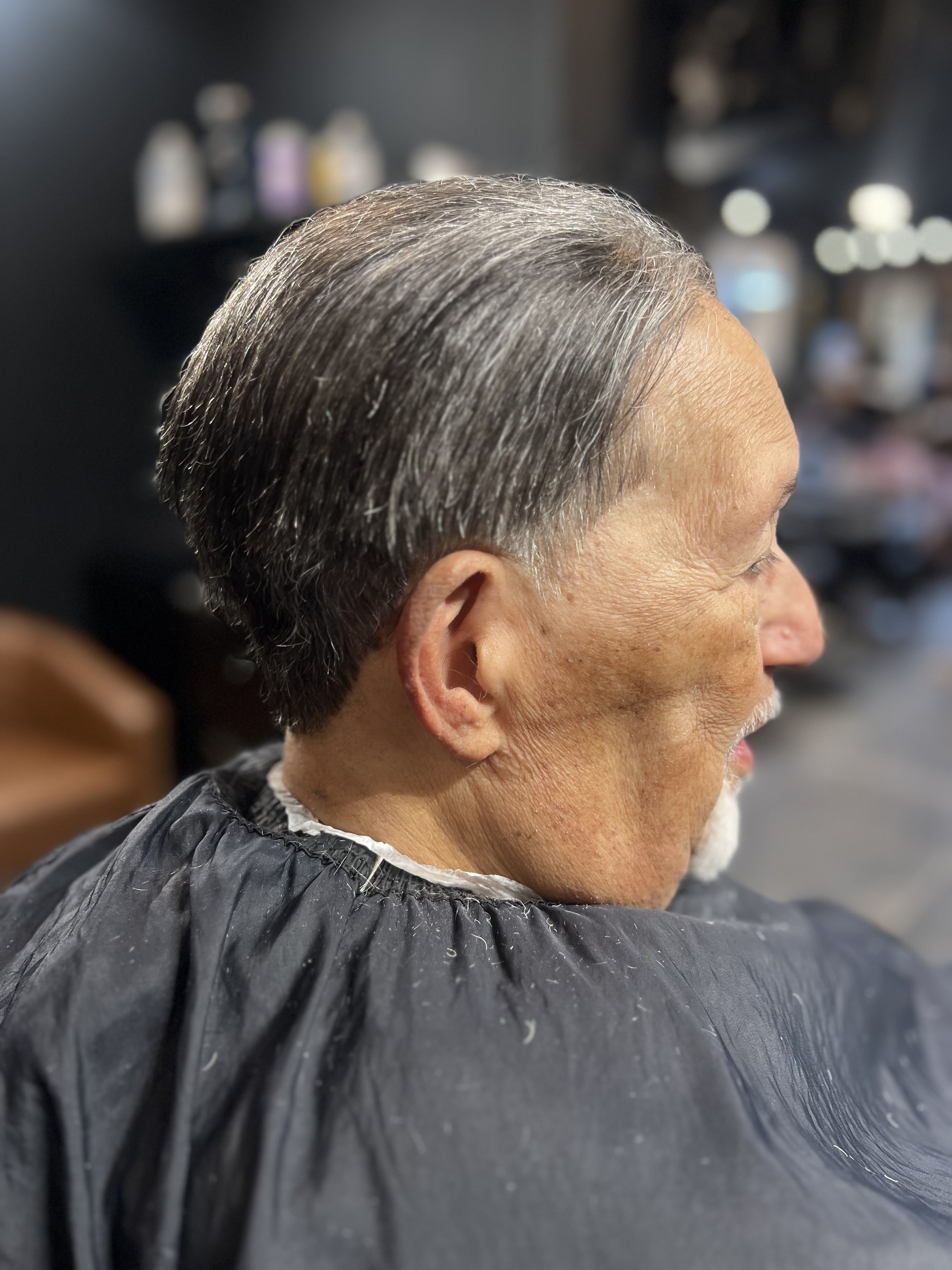Side view of an elderly man with short gray and black hair, sitting in a salon with a black cape around his neck.