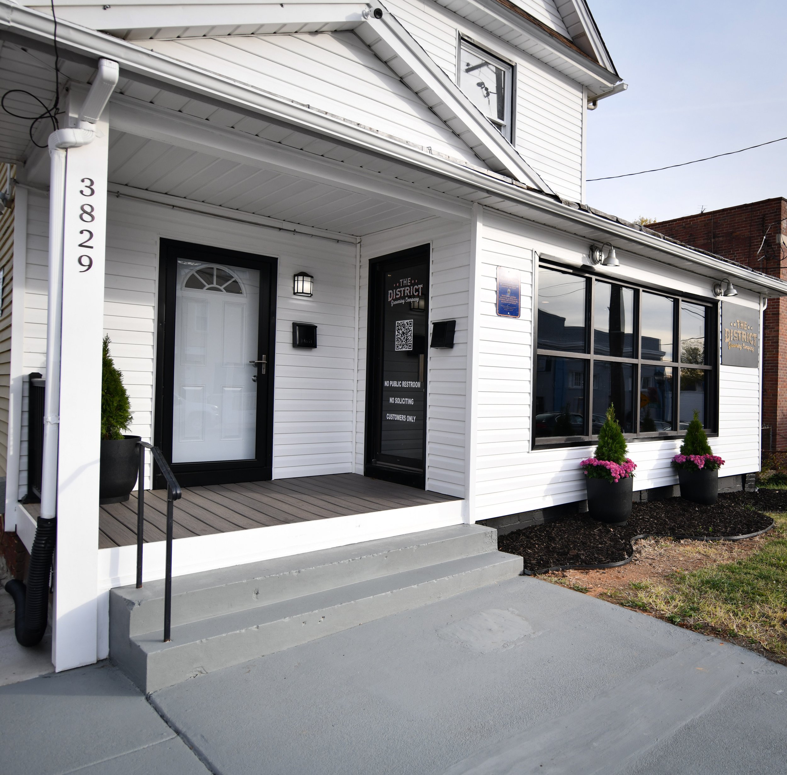 White building with black trim featuring a front entrance with steps and a flat roof. The door displays a sign reading 'The District.' Large windows are adorned with potted plants with pink flowers. The building number 3829 is visible on a post.