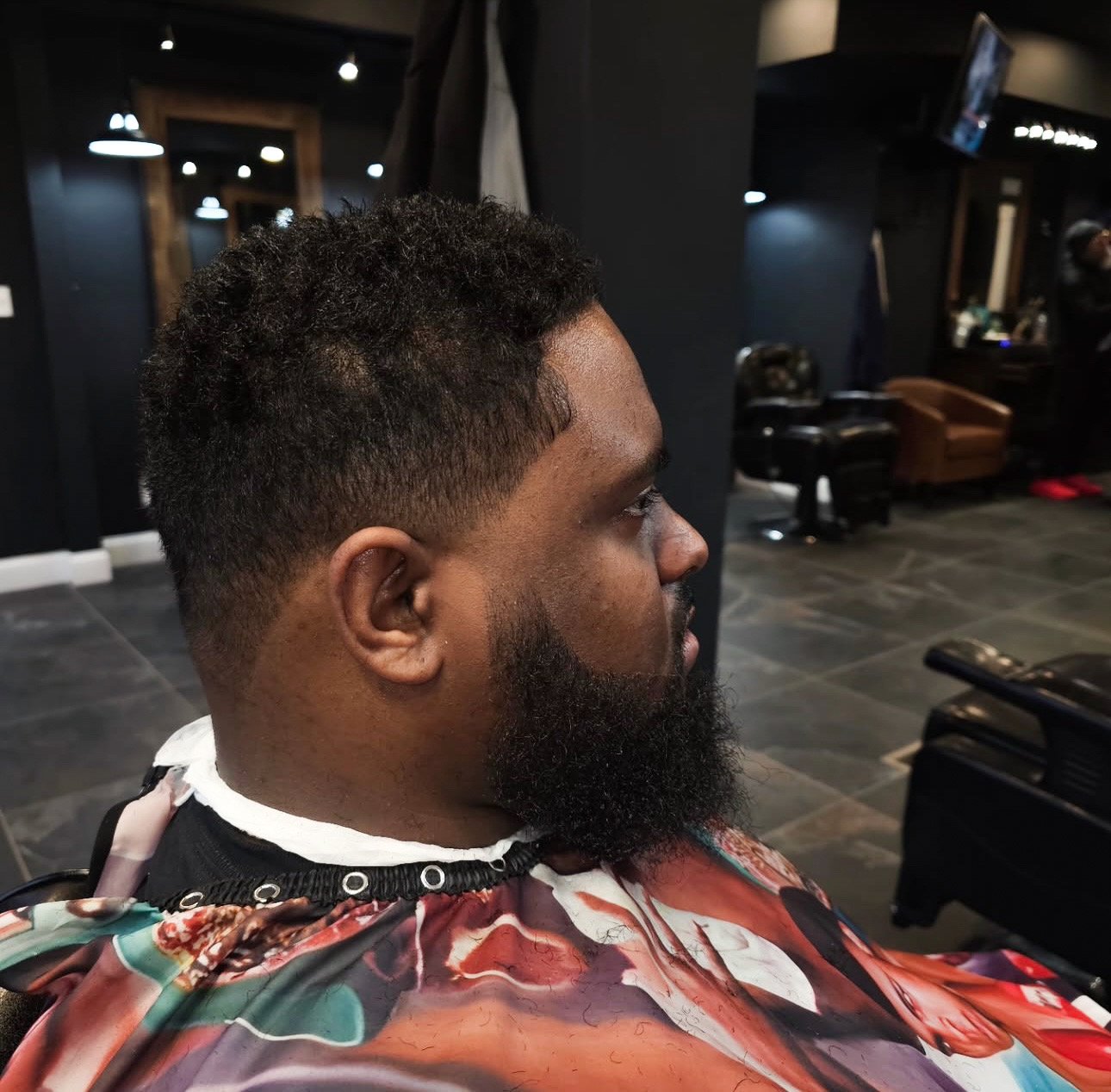 Man sitting in barber shop with a fresh haircut and beard, wearing a colorful barber cape.