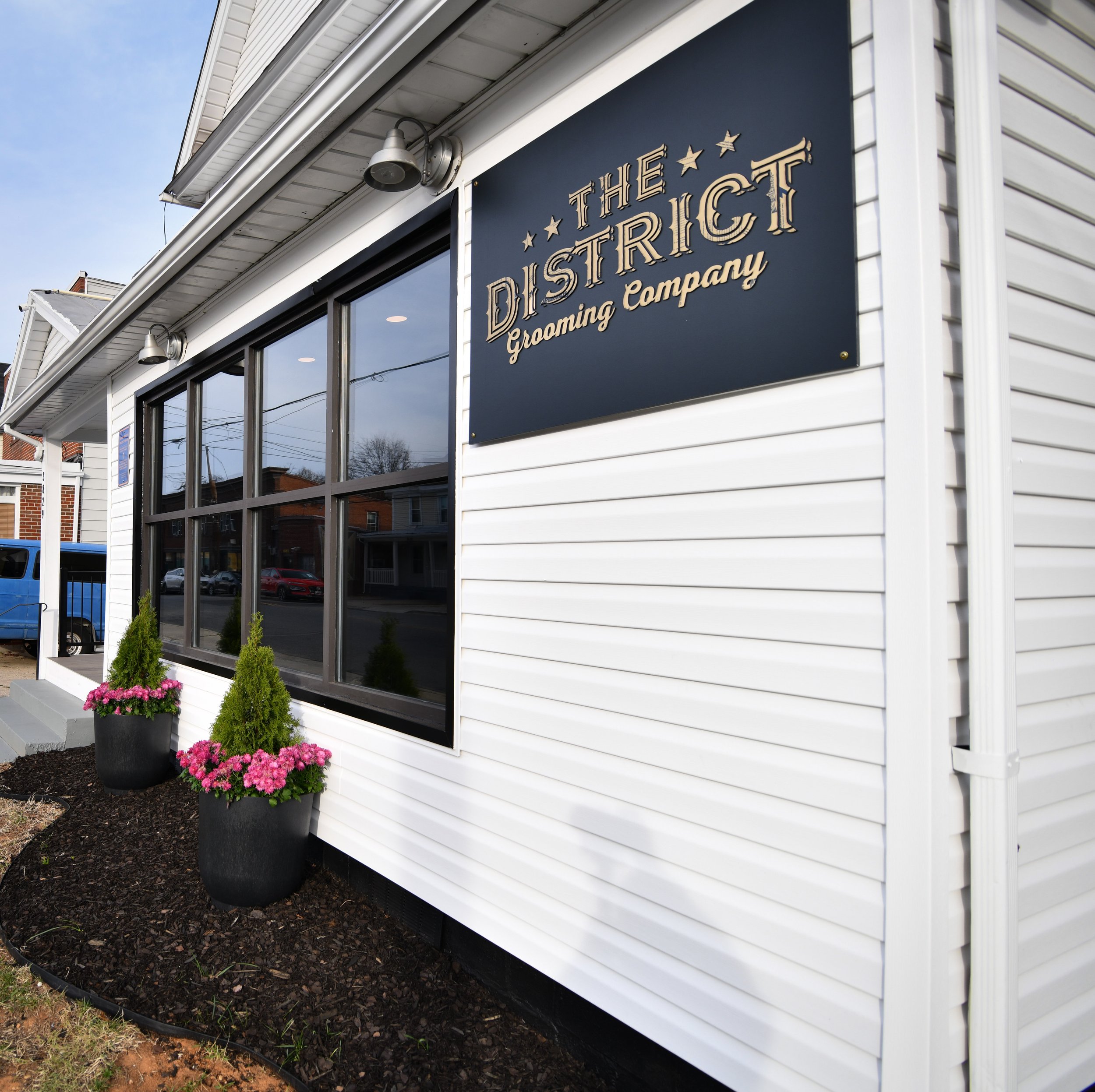 Exterior of The District Grooming Company with a white facade, large windows, and potted plants.