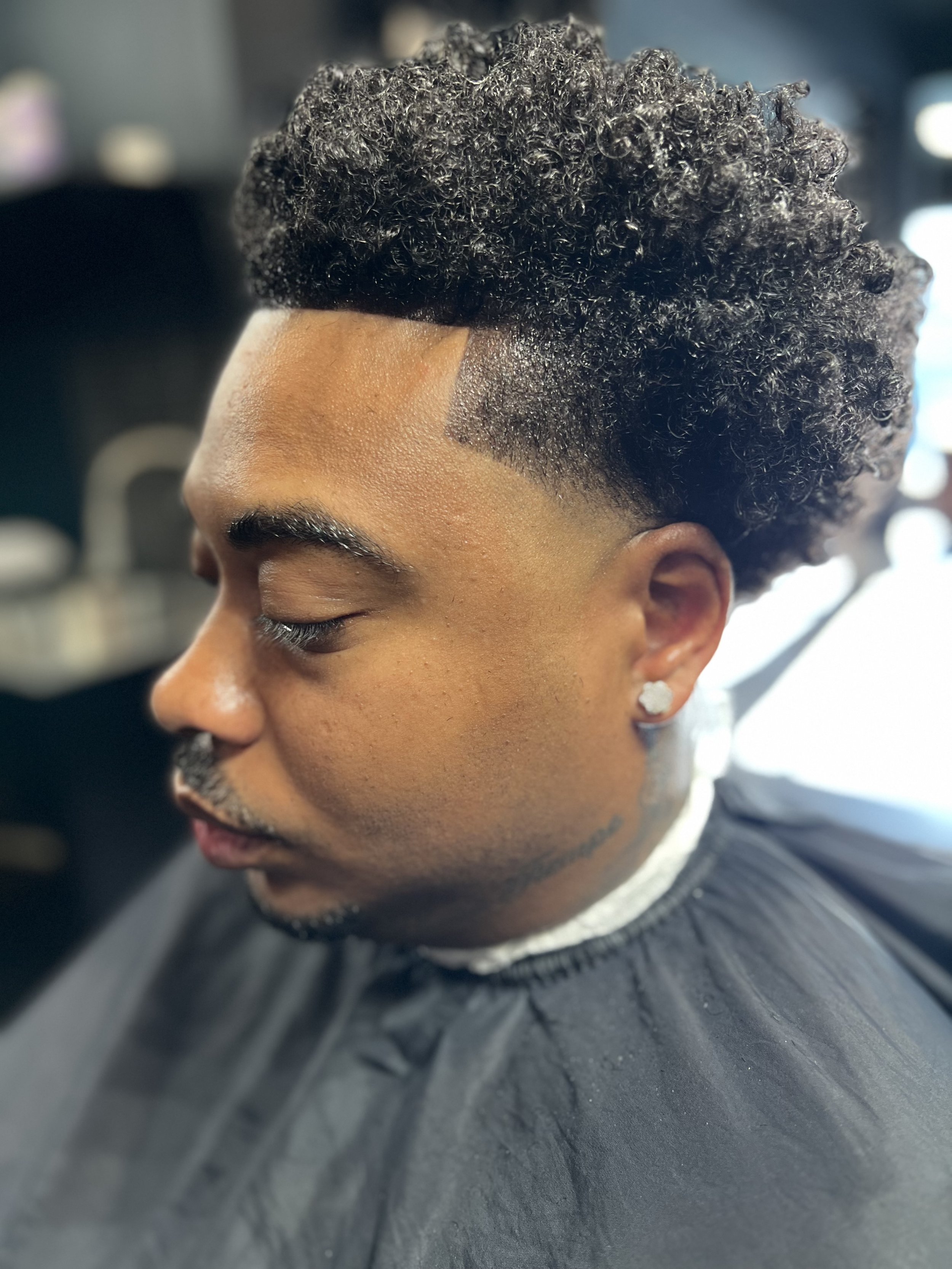 Close-up side profile of a young man with a high fade haircut with curly hair on top, wearing a black barber cape, silver earring, and having his eyes closed in a barbershop setting.