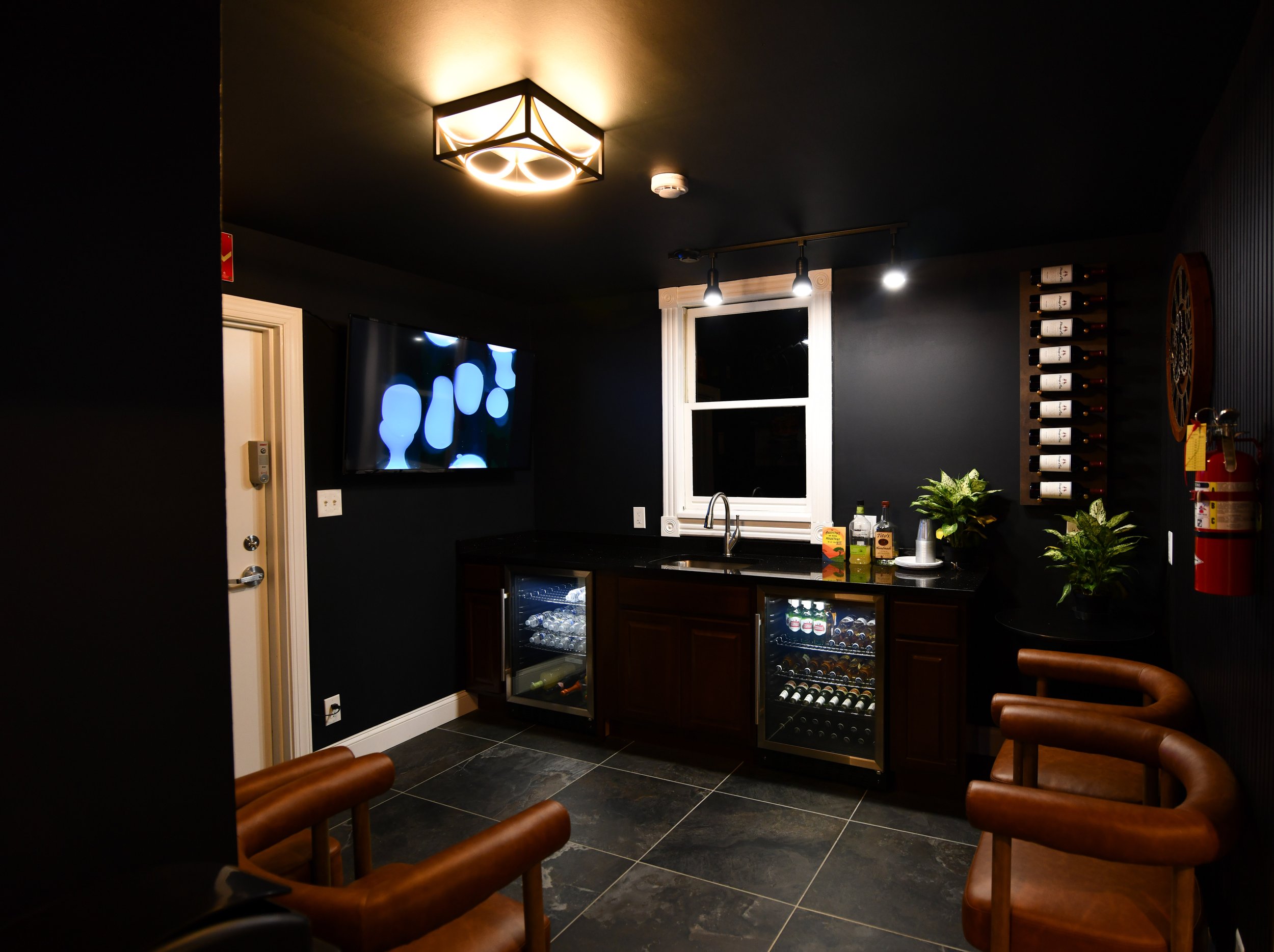 A modern dark-colored room with a bar area, featuring bar stools, a counter with a sink and bottles, a wall-mounted TV, and a wine rack. Drinks and snacks are displayed on the countertop, surrounded by plants and mood lighting.