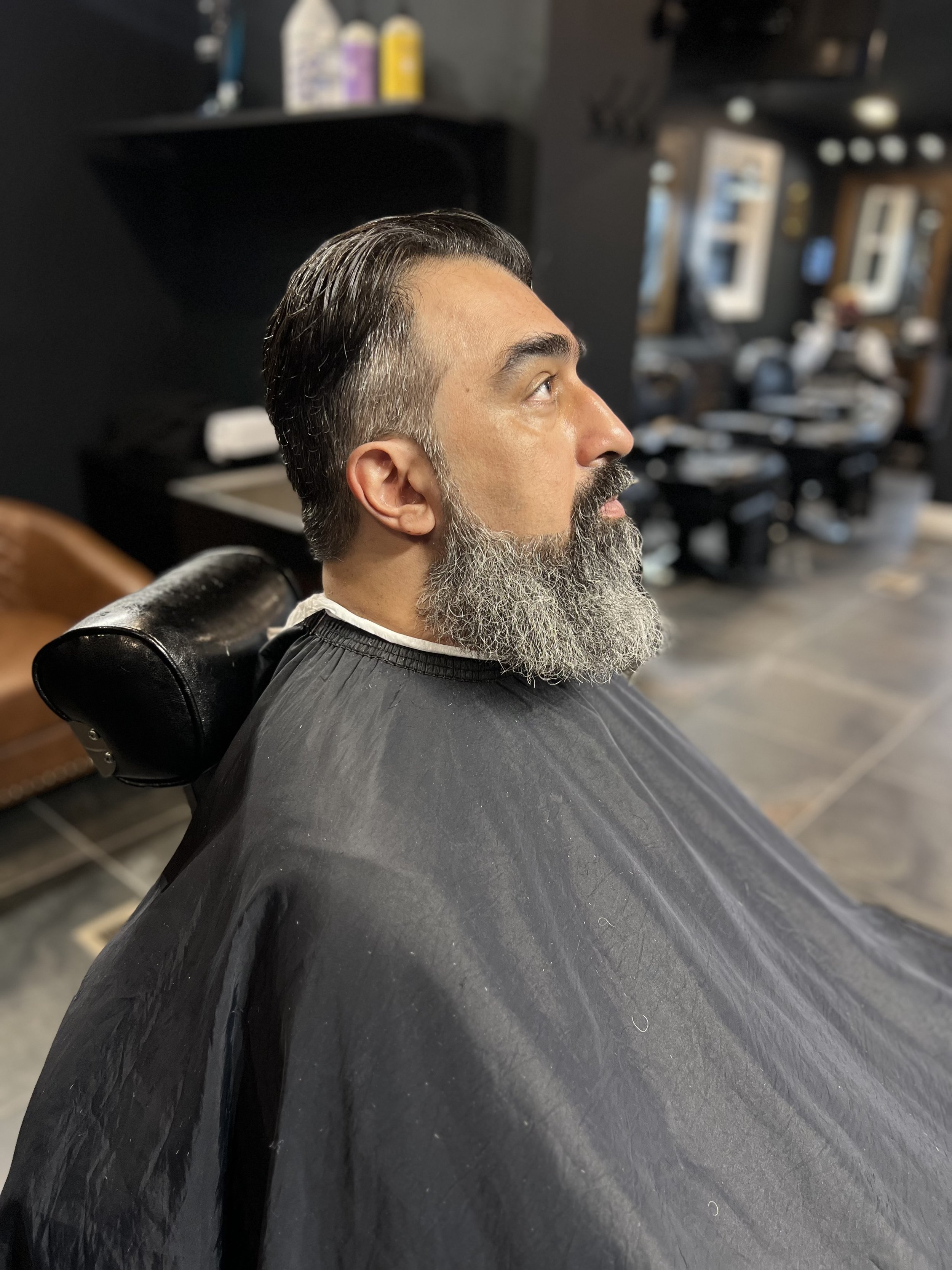 Man with gray beard and slicked-back dark hair sitting in a barber chair at a hair salon, wearing a black cape.
