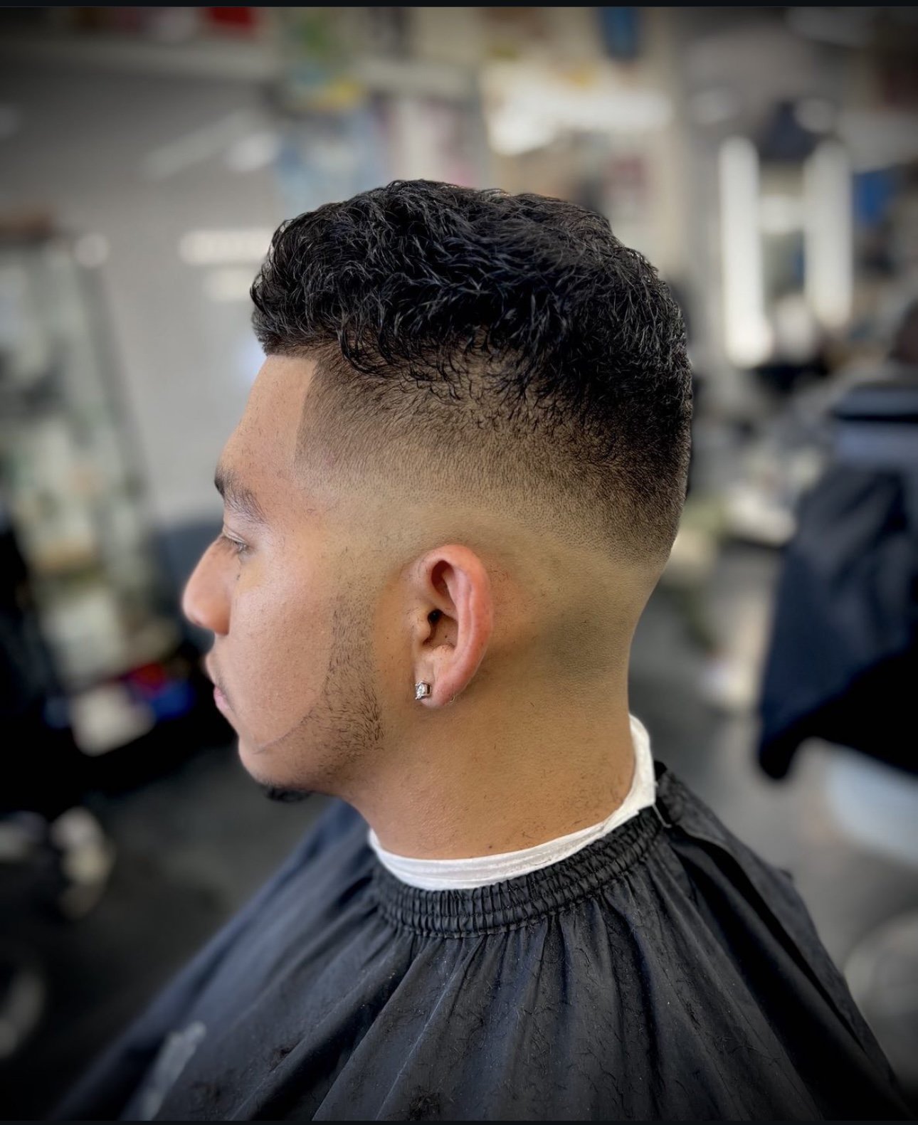 Side view of a young man with a fresh skin fade haircut, dark curly hair on top, and an earring in his ear, sitting in a barbershop.