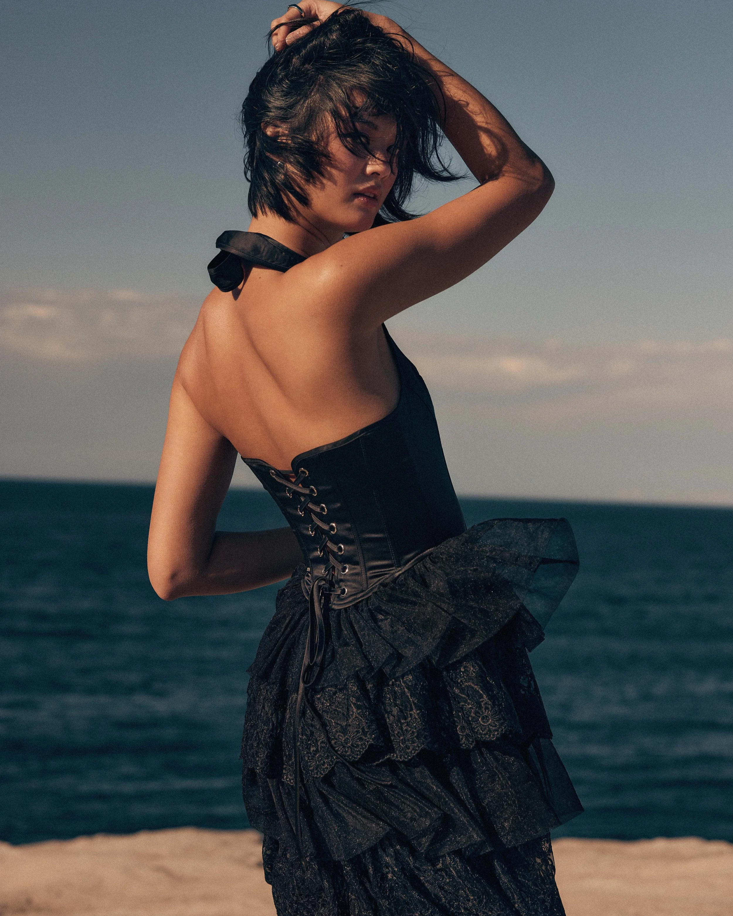A woman with short black hair, wearing a black corset dress with lace details and ruffles, standing by the ocean on a sandy beach during sunset, with her arm raised and hand touching her hair.