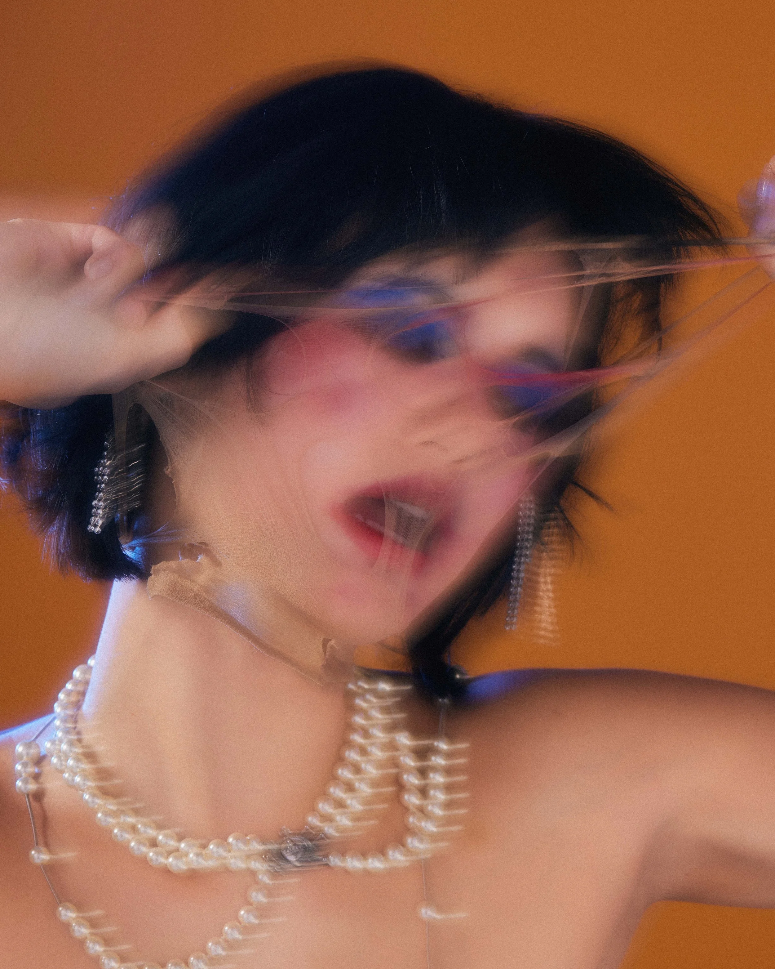 A woman with short black hair, wearing pearl necklaces and earrings, appears to be moving quickly or in motion, creating a blurred effect on her face and jewelry, with an orange background.