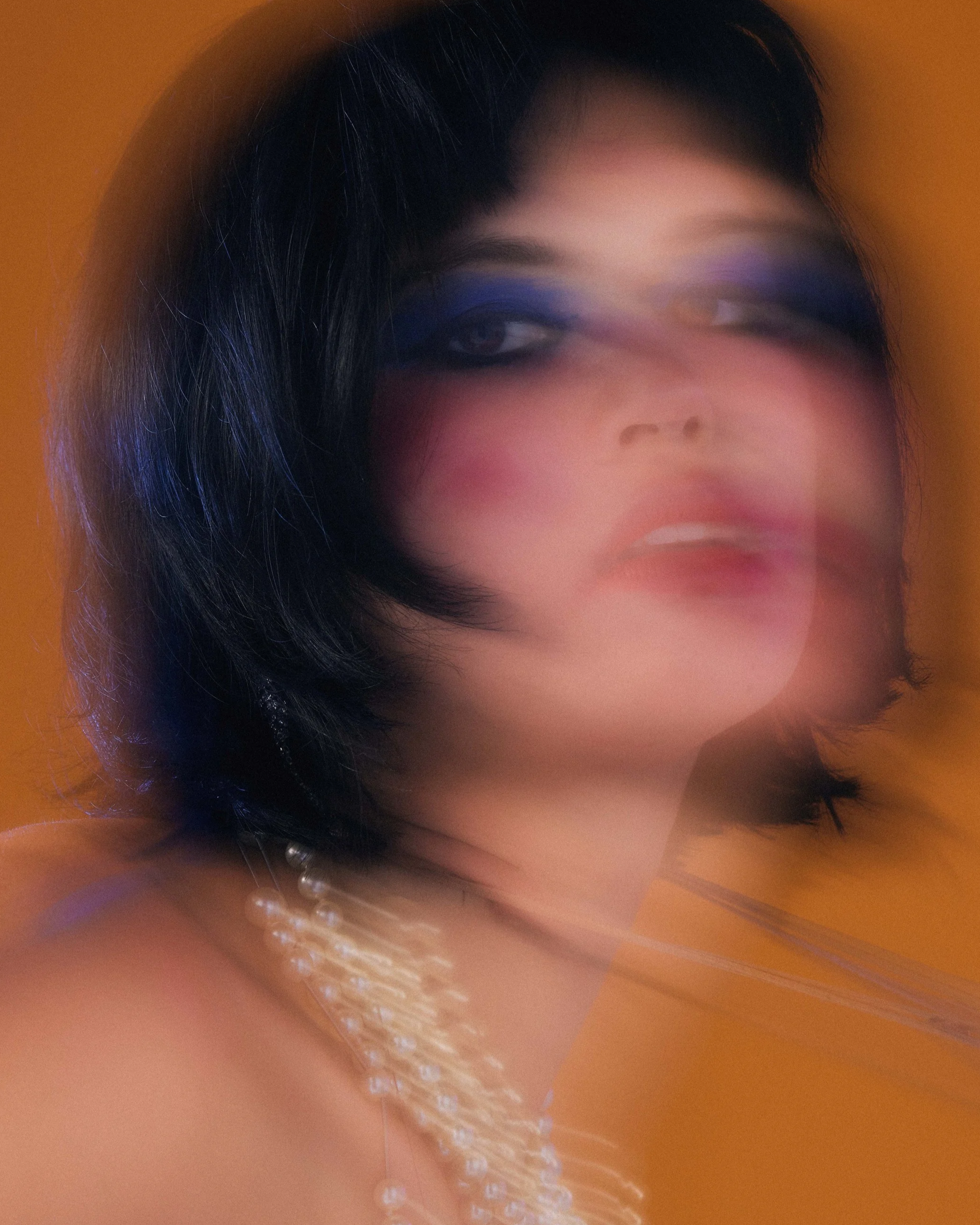 Artist with short black hair wearing a pearl necklace and bold makeup, with motion blur creating a surreal, dreamy effect against an orange background.