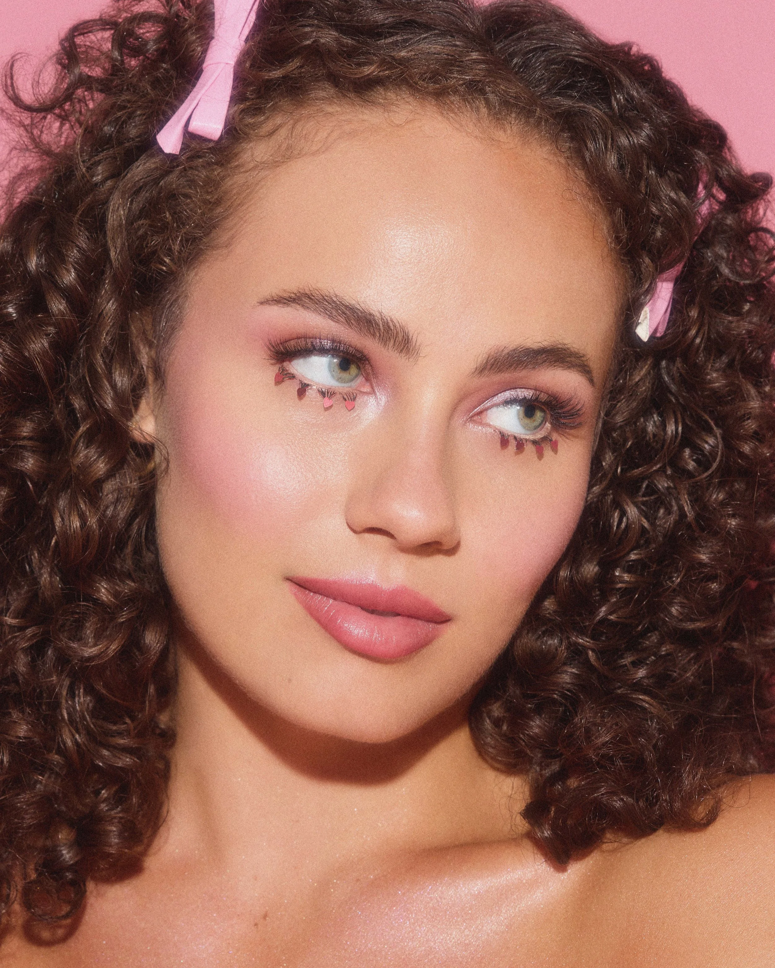 Close-up of a woman with curly brown hair, pink hair clips, and makeup with pink eyeshadow, eyeliner, mascara, and pink heart-shaped decorations under her eyes, against a pink background.