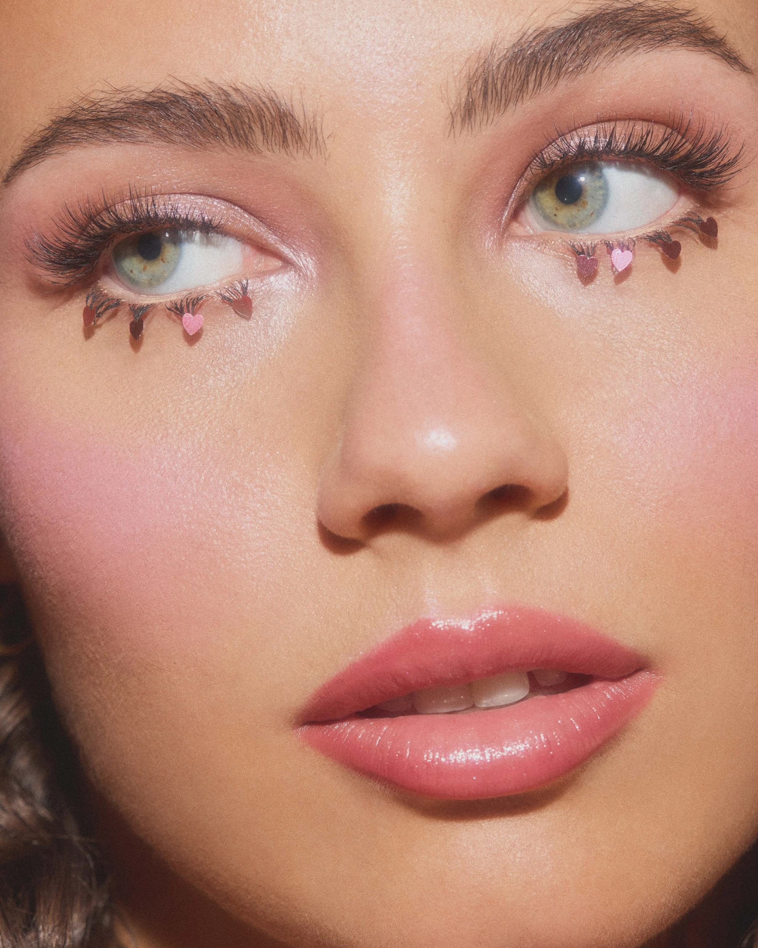 Close-up of a woman's face showing her eyes, nose, and lips. Her eye makeup includes pink eyeshadow and false eyelashes with small heart-shaped decorations hanging from her lower eyelashes.