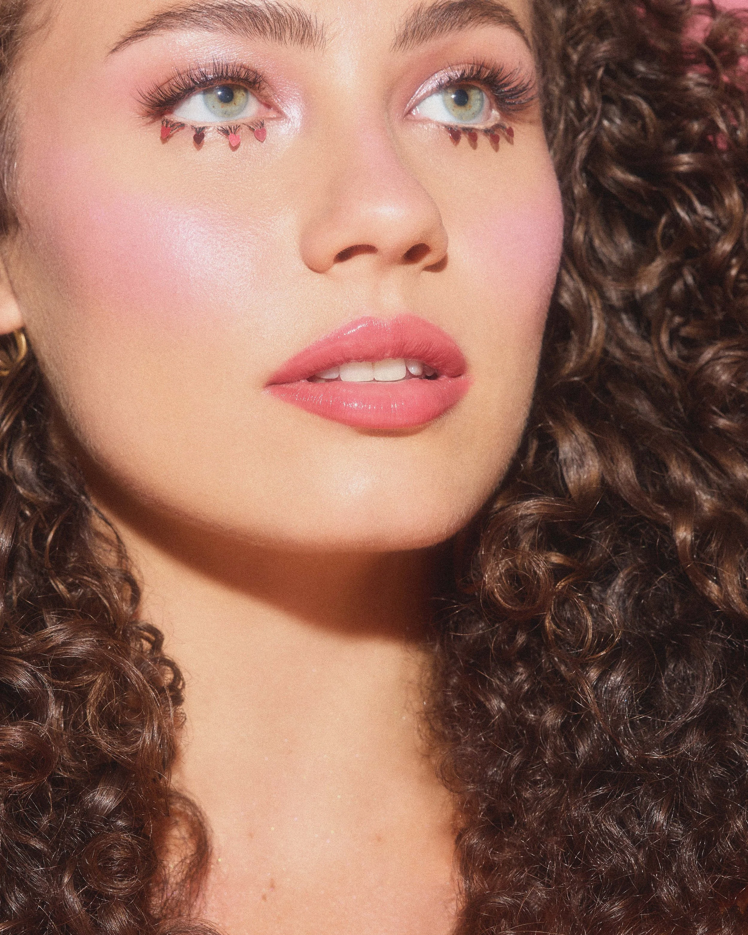 Close-up of a woman with curly hair, makeup, and pink heart teardrop designs under her eyes.