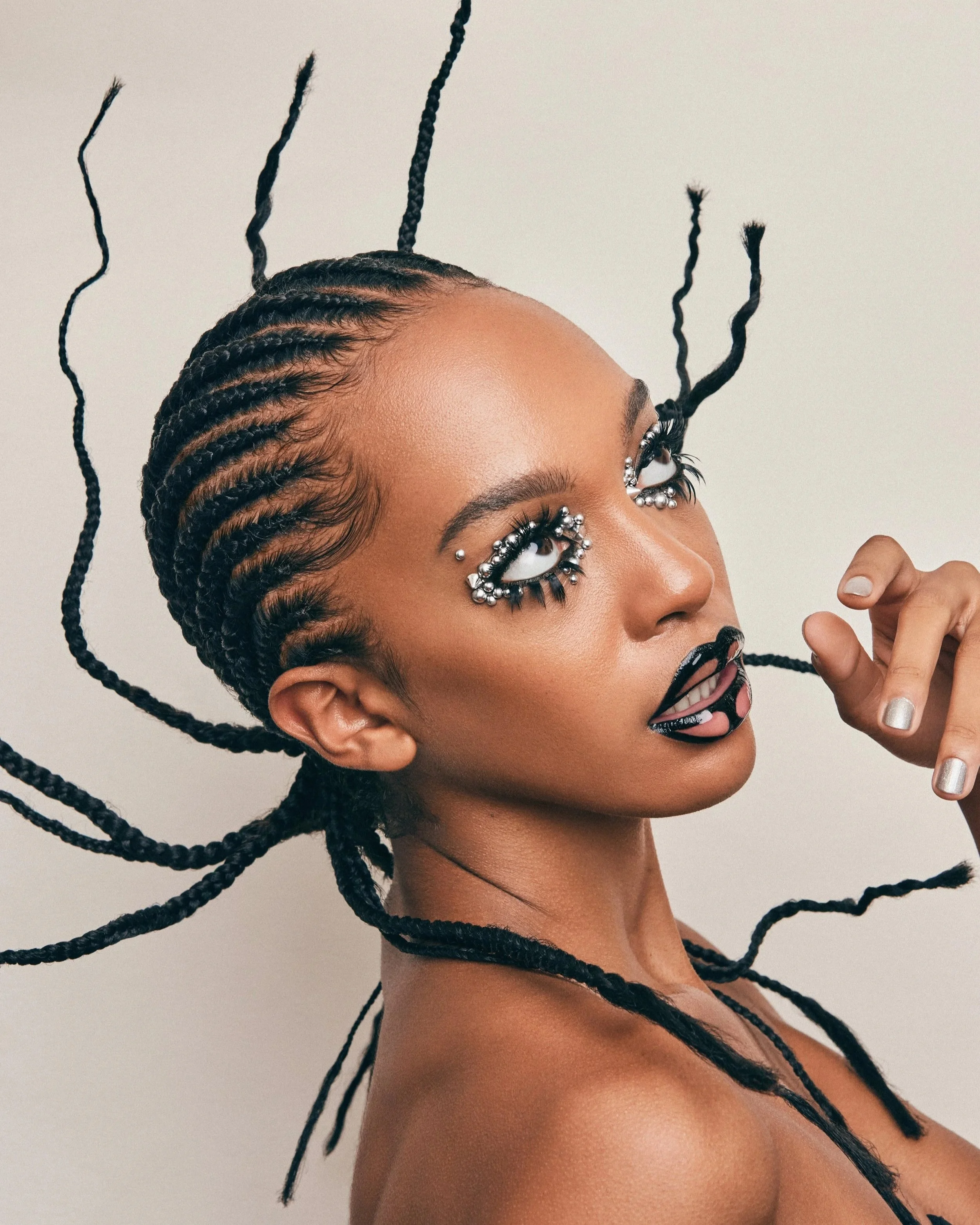 A woman with braided hair and elaborate makeup, including pearls around her eyes and black lipstick, posing against a plain background.