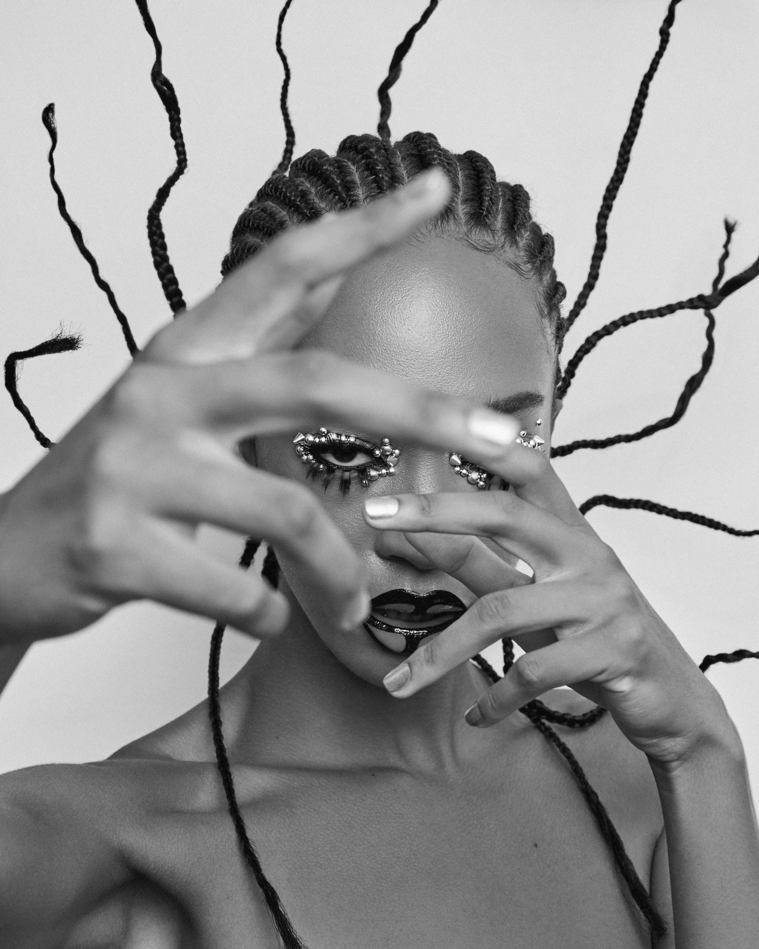 Black and white portrait of a woman with braided hair and dramatic makeup, covering her face with her hands, in front of a plain background.