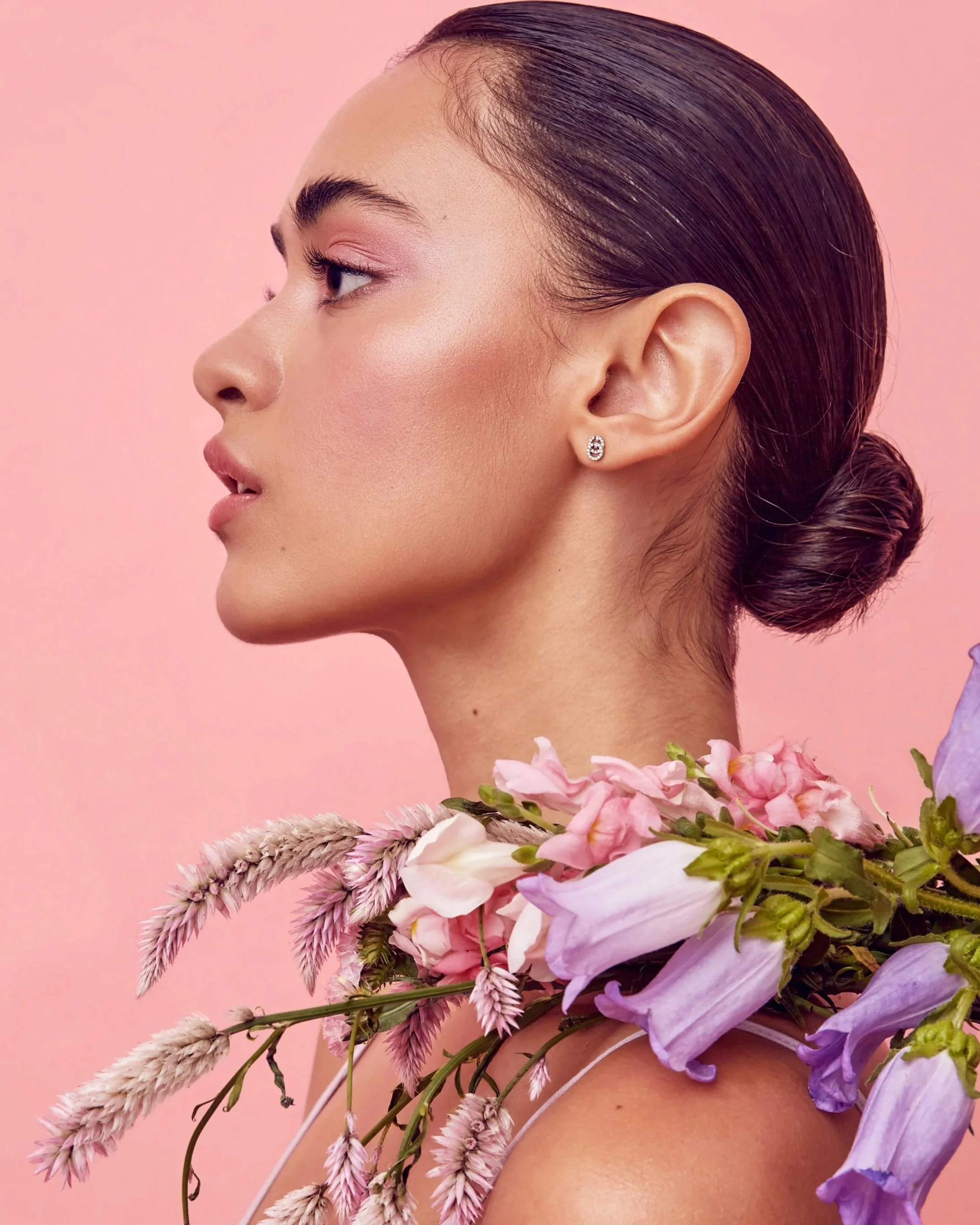 Profile of a woman with dark hair in a bun, pink makeup, wearing a pearl earring, with a flower necklace of pink, purple, and white flowers against a pink background.
