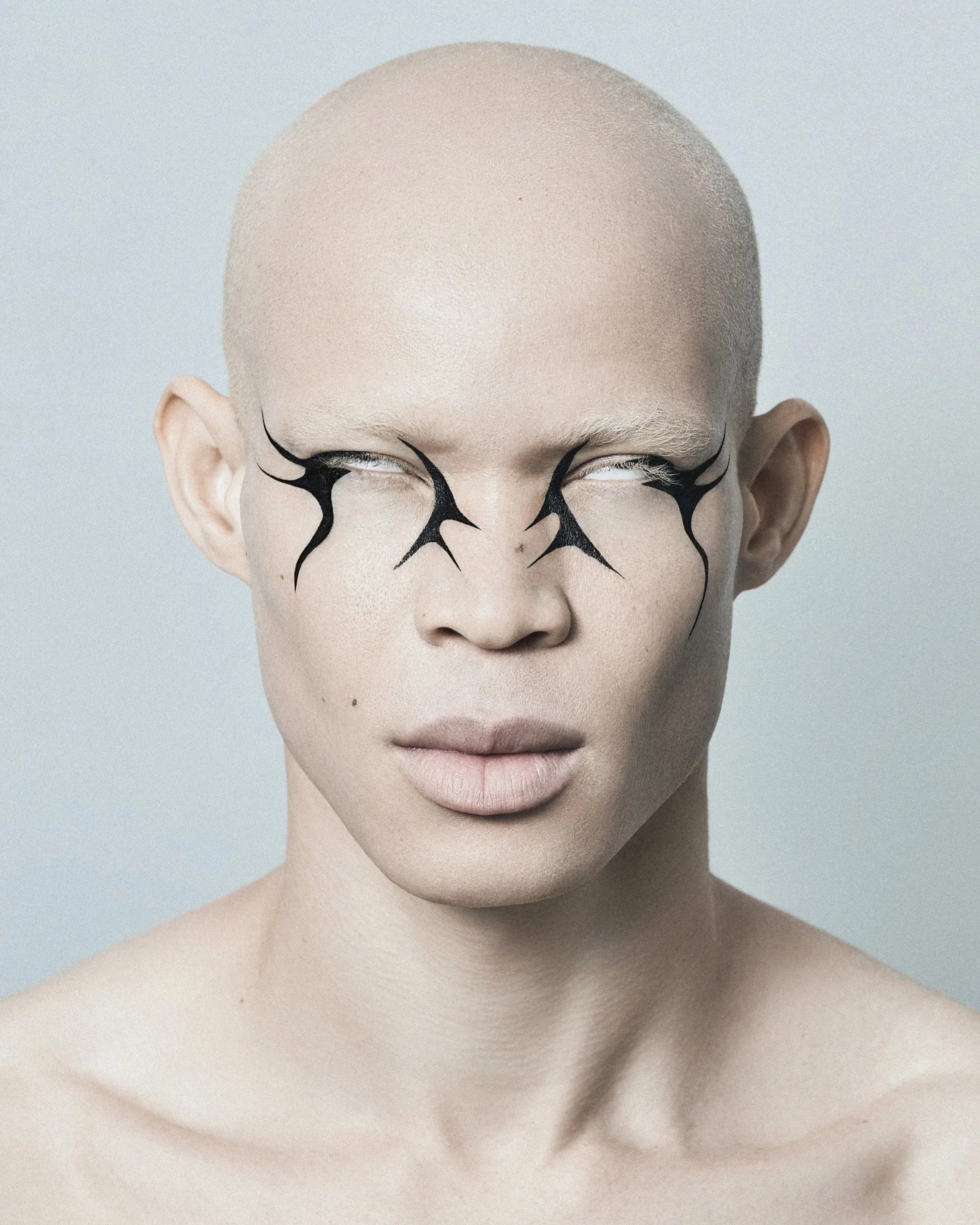 A person with a shaved head and white skin, wearing black abstract eye makeup that extends outward from the eyes.