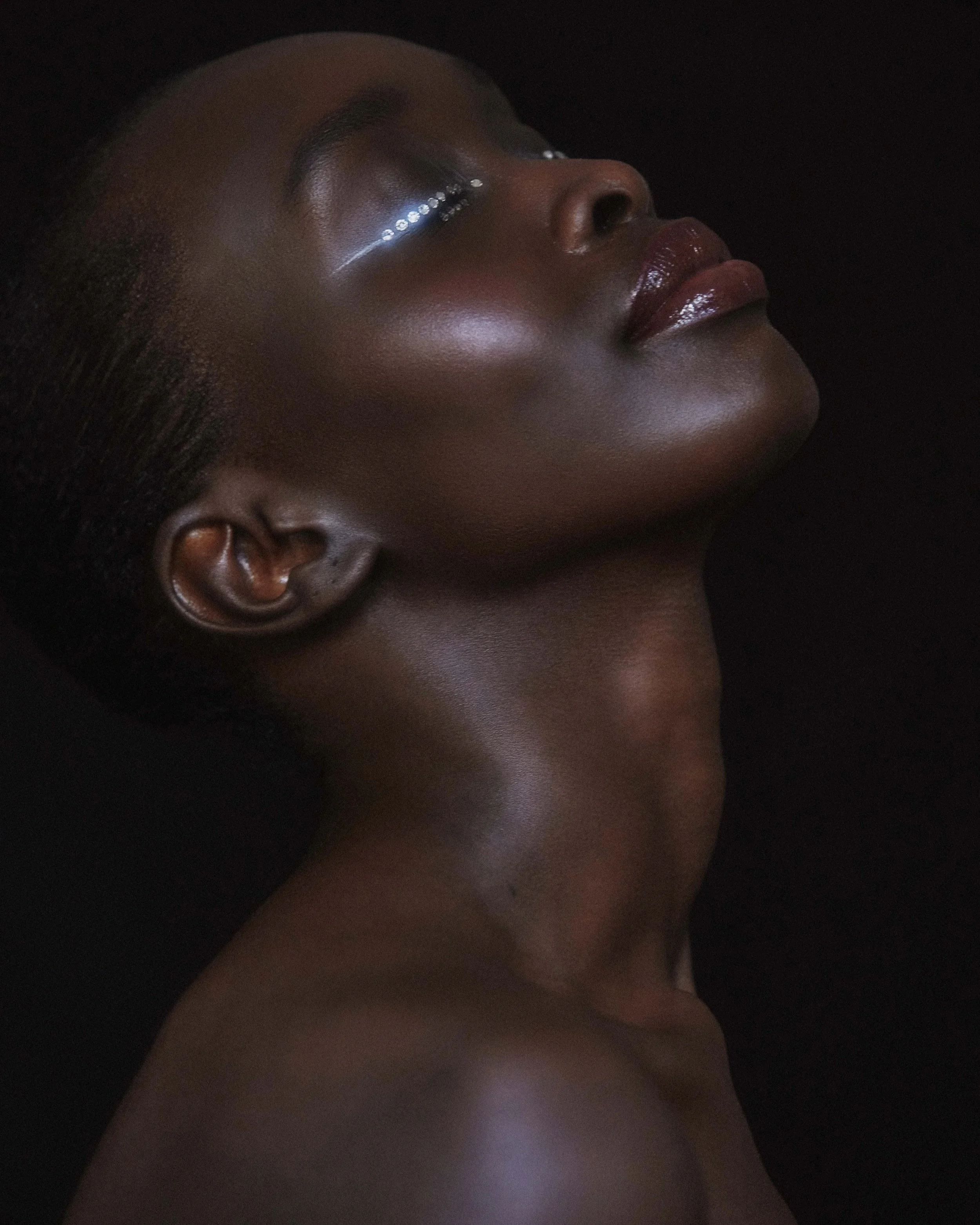 Close-up of a woman with dark skin, her eyes closed, showing glossy lips and eye makeup, against a black background.