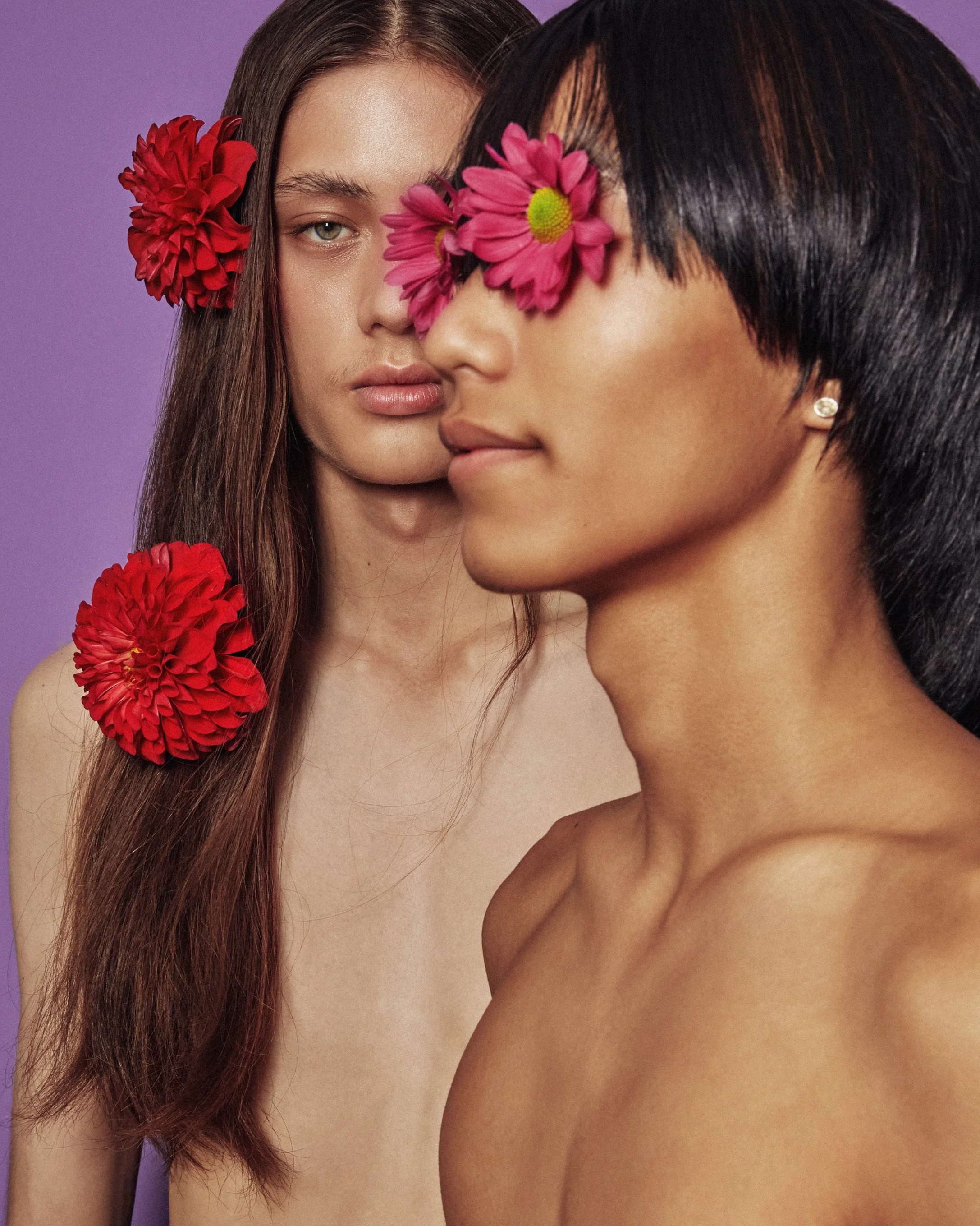 Two topless women, one with long brown hair and a red flower in her hair, the other with short black hair and a pink flower, covering their eyes with flowers, against a purple background.