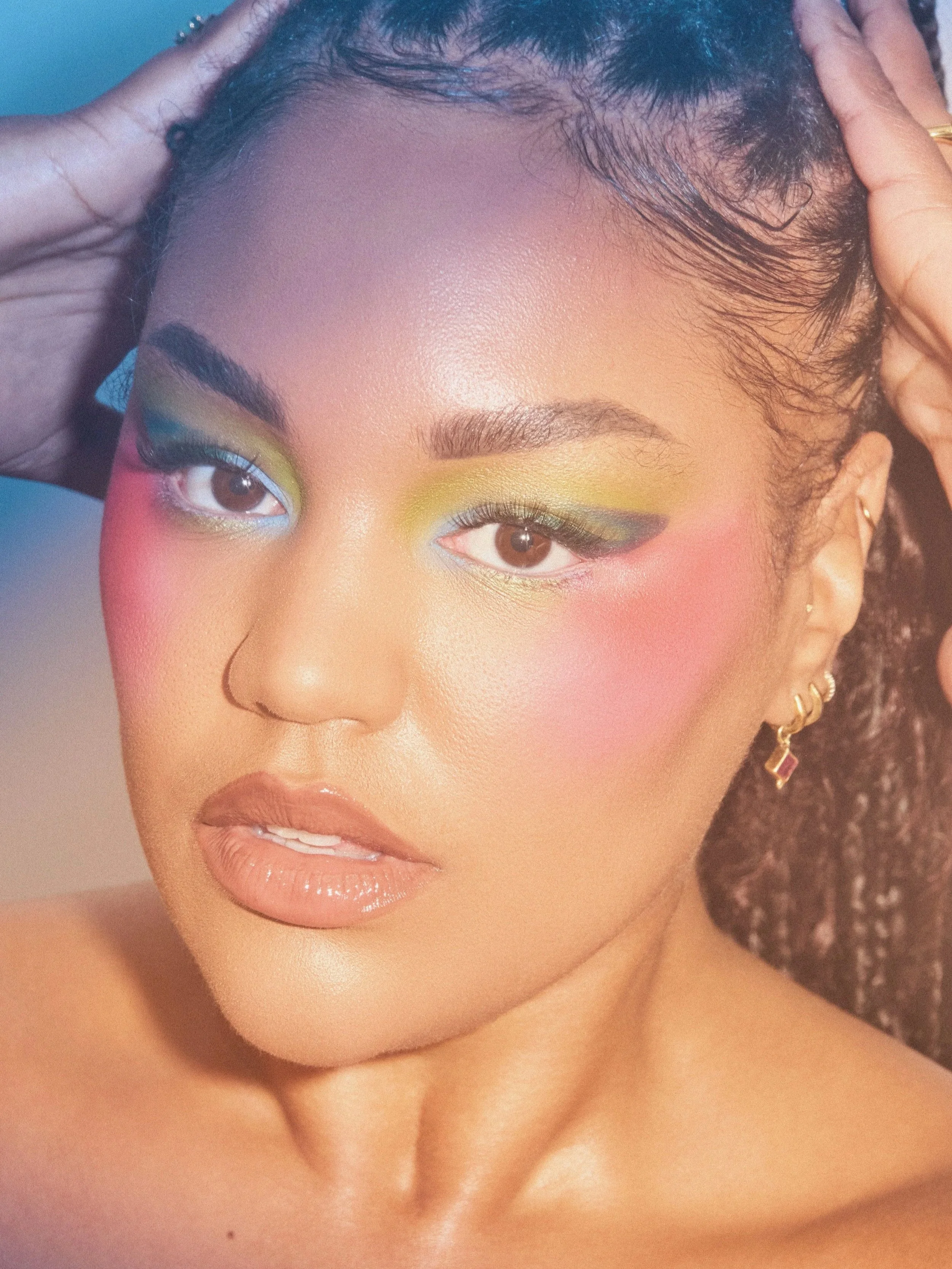 Close-up of a woman with colorful makeup including yellow, green, and pink eyeshadow, glossy lips, and wearing gold jewelry.
