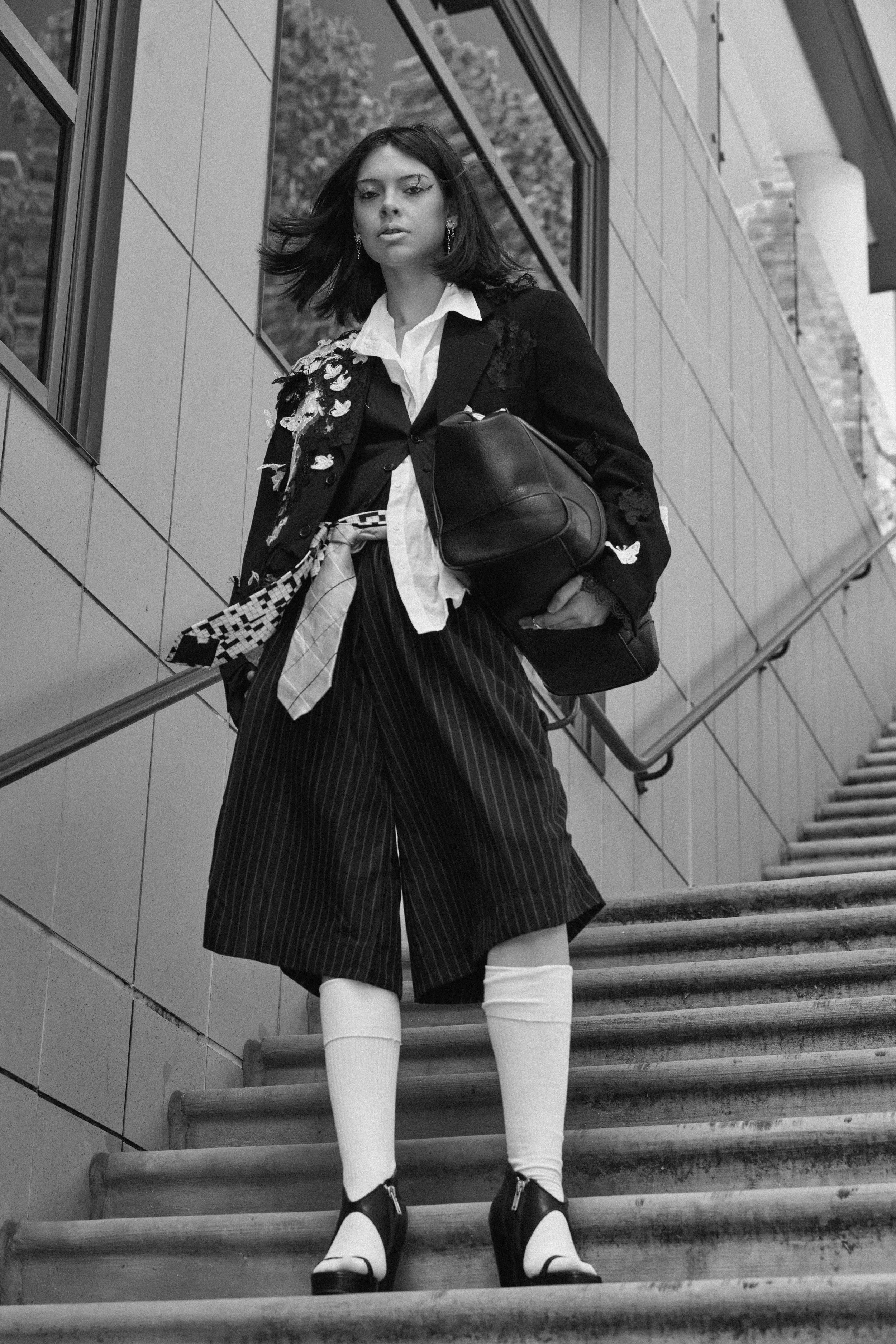 A woman with dark, shoulder-length hair stands on outdoor stairs, looking at the camera. She wears a patterned blazer, a white shirt, pinstriped culottes, knee-high socks, and high-heeled shoes. She holds a black bag and is in front of a modern build