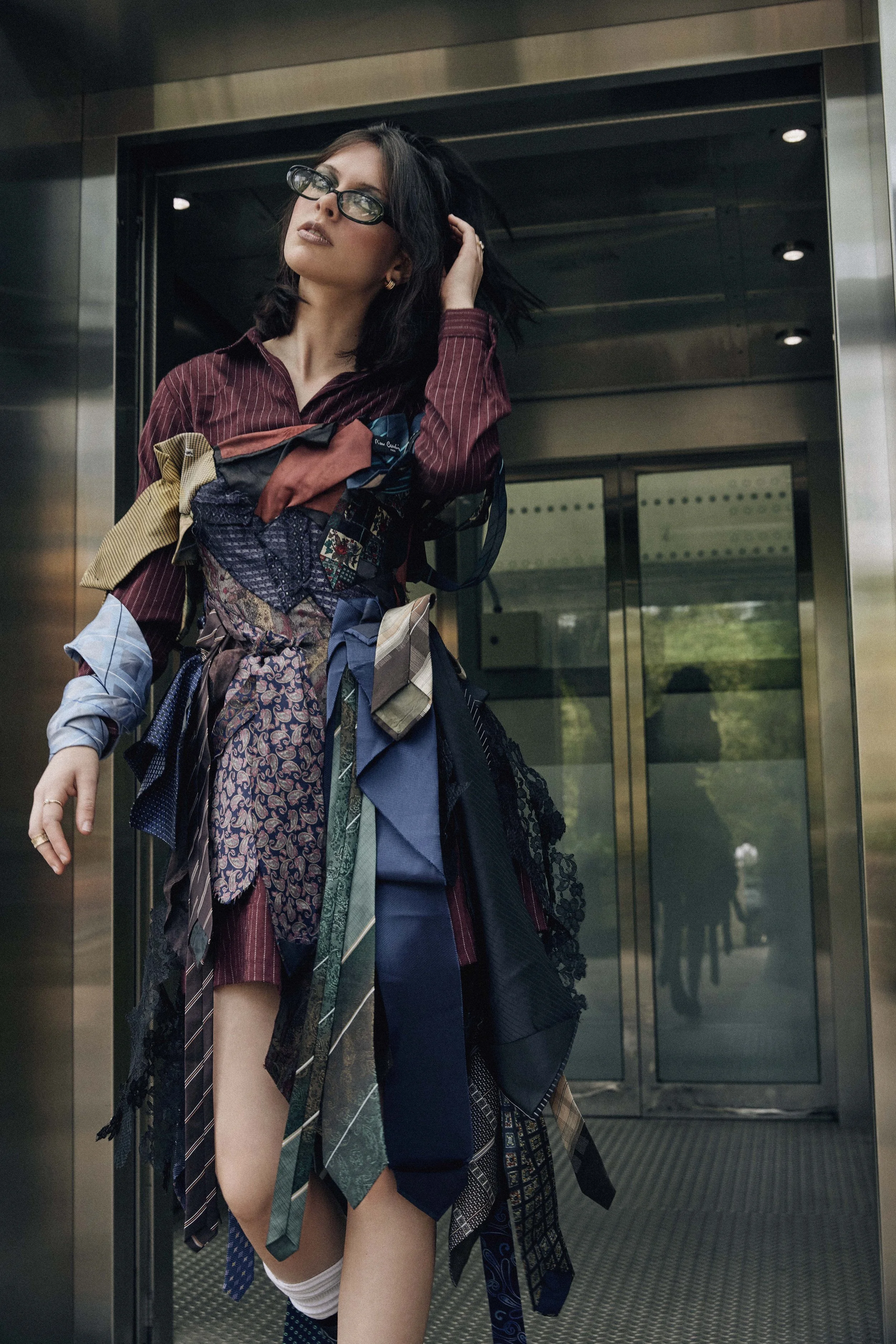Woman with black hair and glasses wearing a dress made of various fabric swatches, standing in front of an elevator.