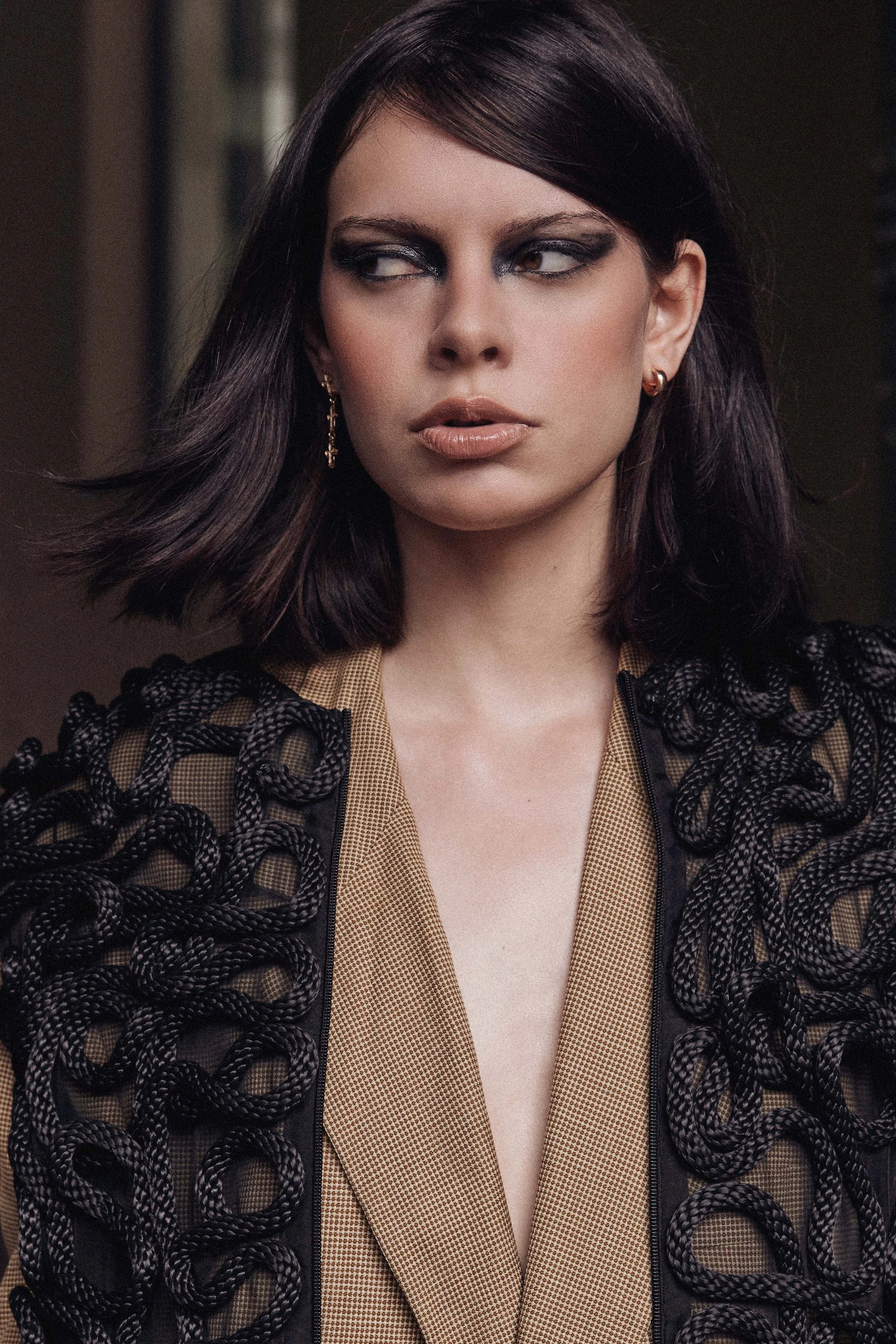 A woman with dark brown hair wearing eye makeup, earrings, a beige shirt, and a dark, intricately woven garment.