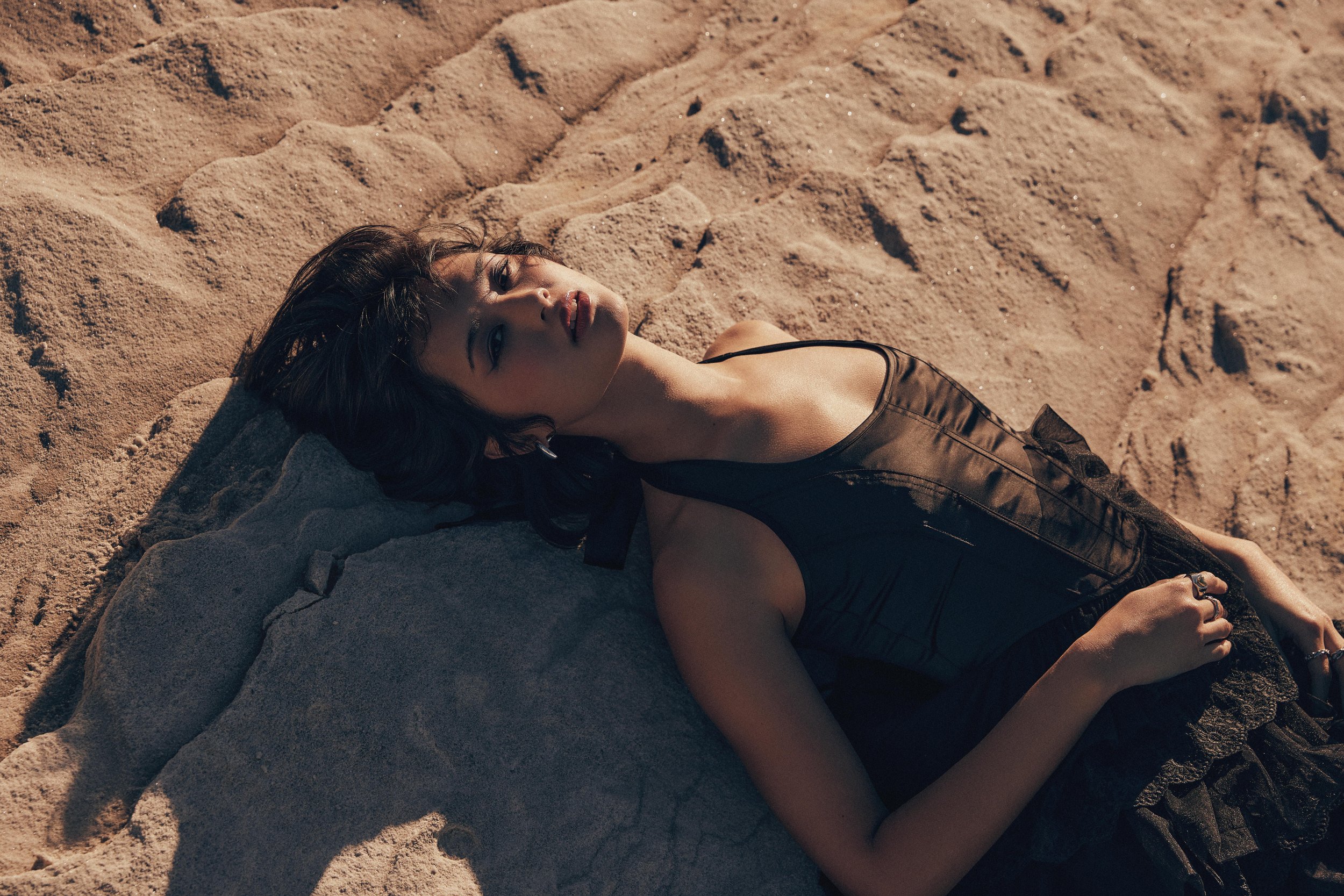 A woman lying on a rock at the beach, wearing a black dress, with sand around her.