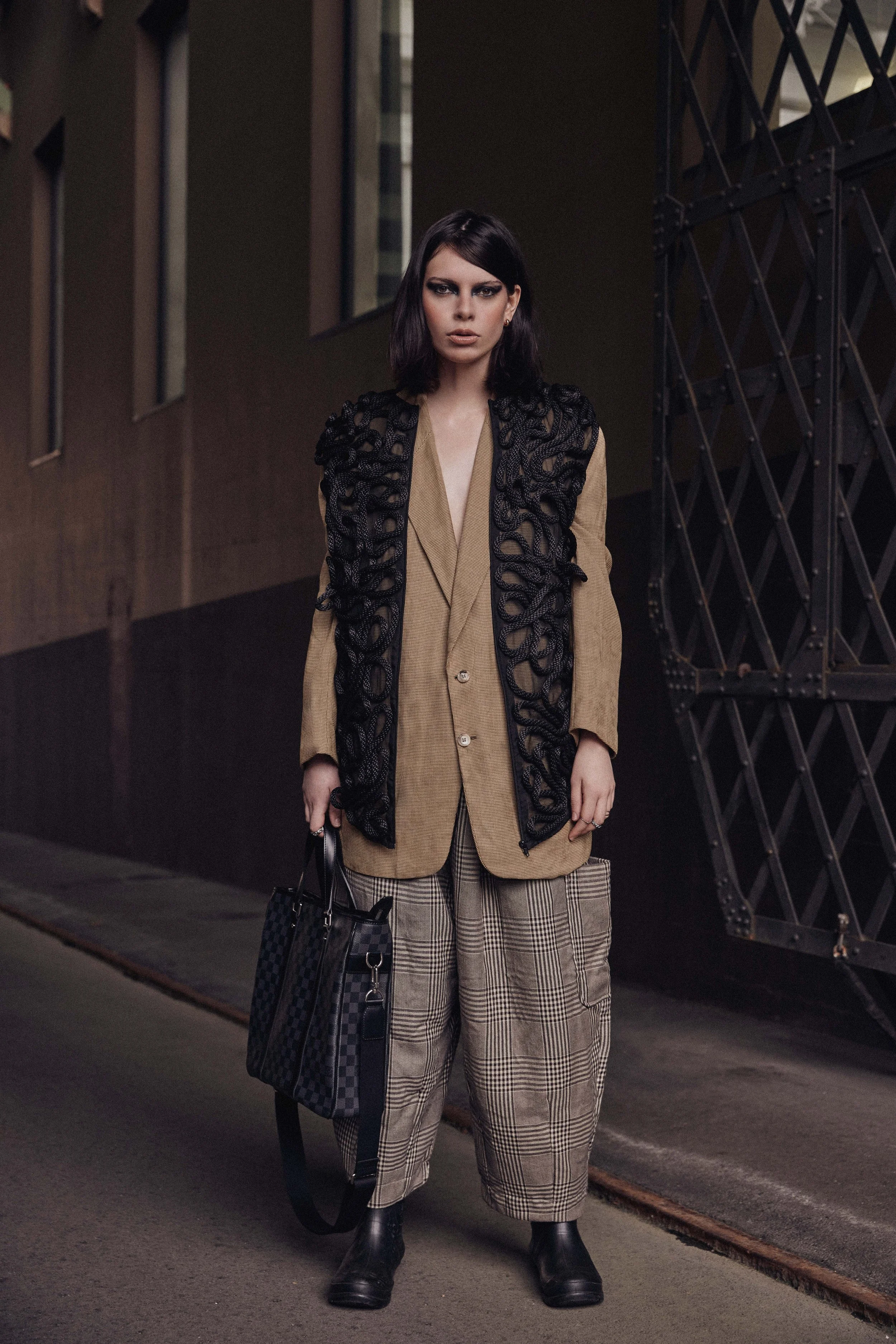A woman with dark hair dressed in a beige blazer, black lace vest, plaid pants, and black boots standing on a city street.