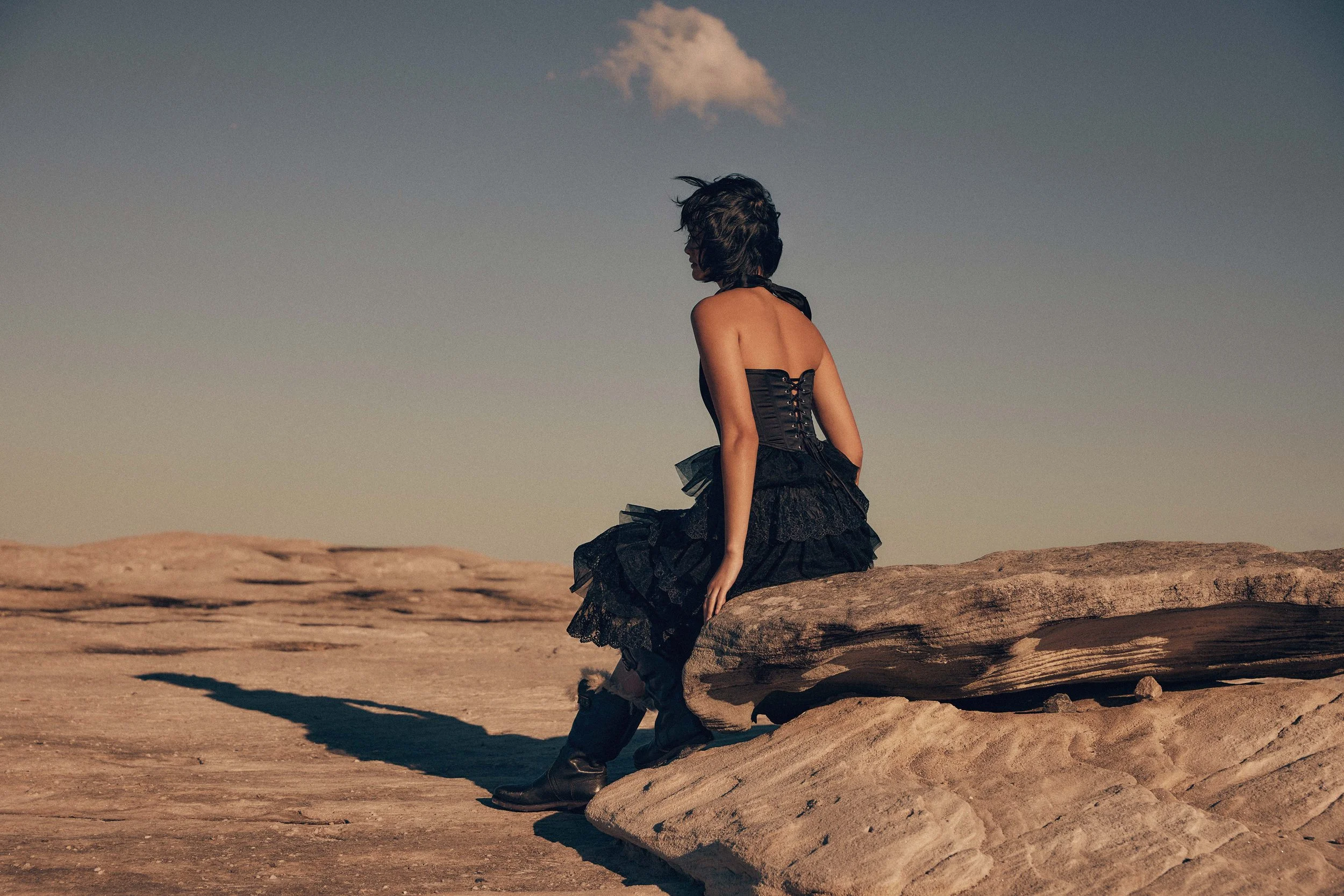 A person dressed in a black lace dress and black boots, sitting on a large rock in a desert landscape with a clear sky.