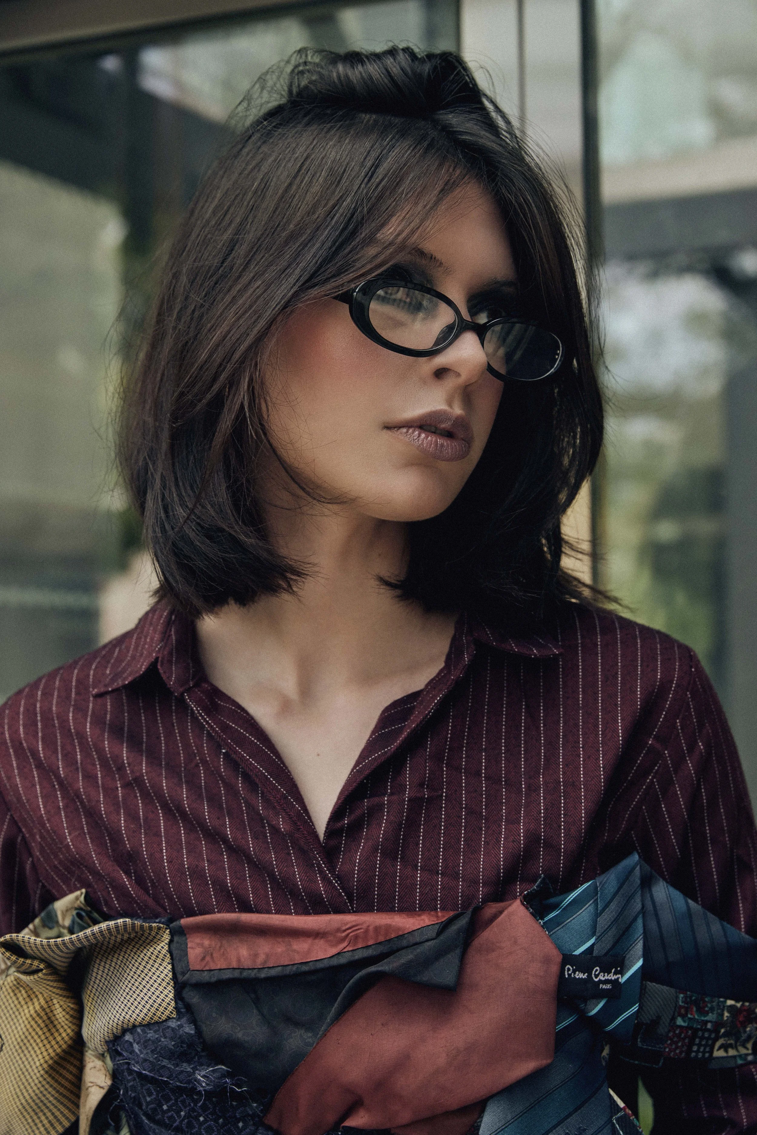 A woman with shoulder-length dark hair wearing black glasses and a maroon pinstripe shirt, holding a colorful patchwork scarf or clothing item, standing outside near a glass building.