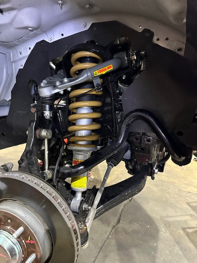 A close-up view of a 4WD Tough Dog suspension system, featuring a coilover shock absorber and brake disc, with a yellow label visible on the shock.