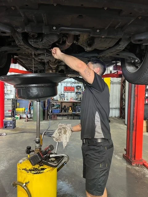 Mechanic changing oil in a car at an auto repair shop, with vehicle lifted on a hydraulic lift and oil draining into a container below.